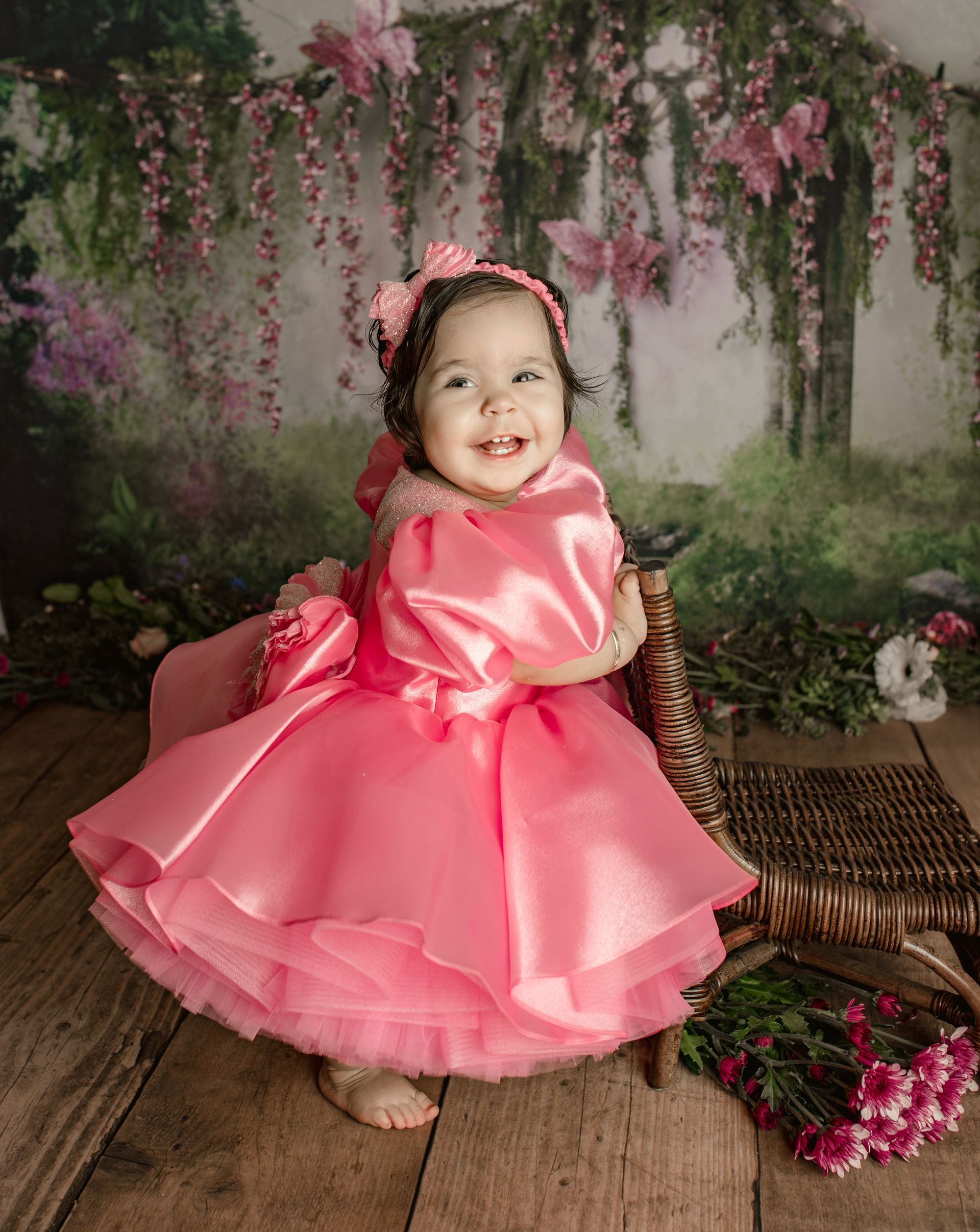 Smiling child in a pink satin dress sitting on a woven bench in a floral garden backdrop. Birthday Session