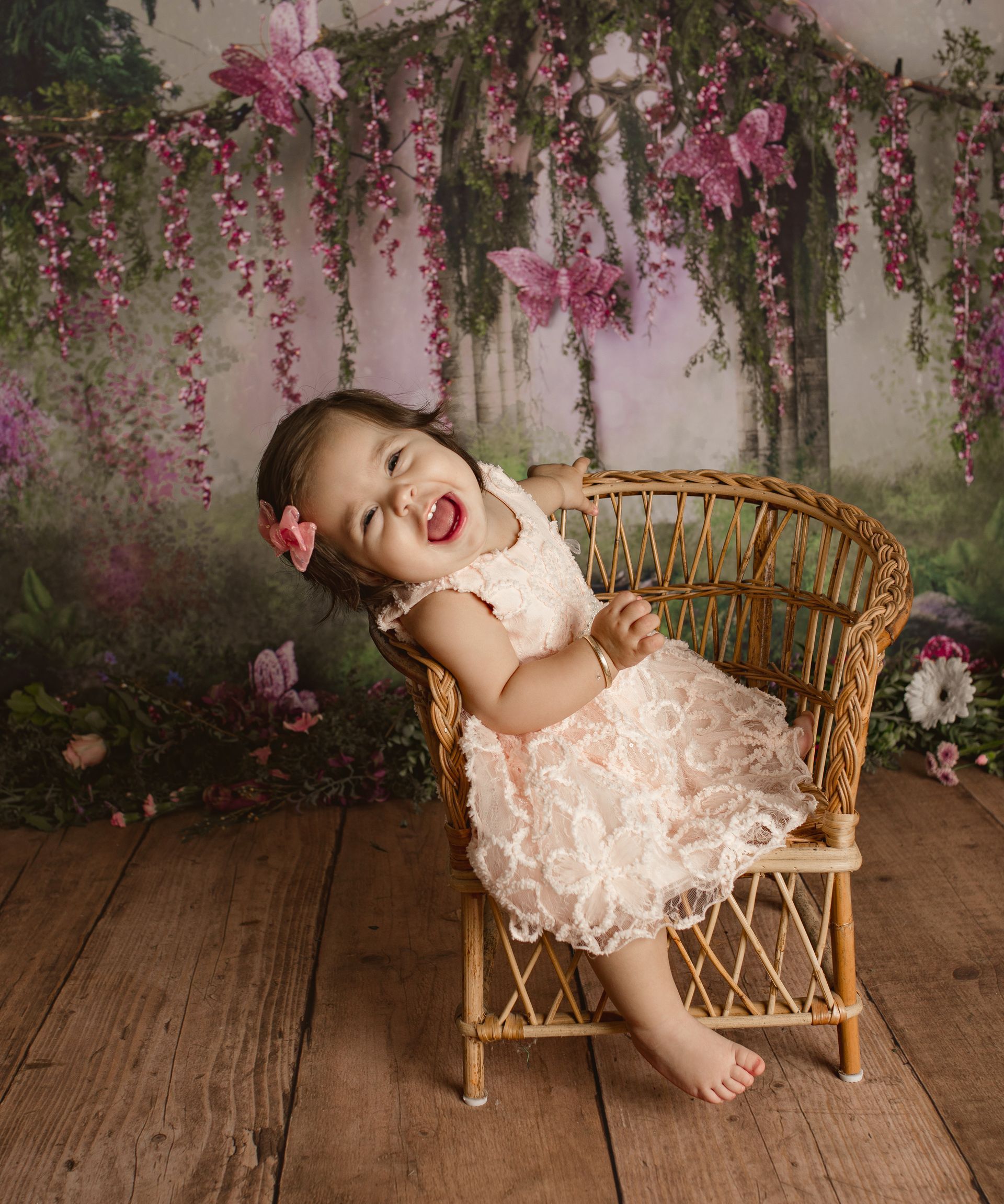 Smiling child in a lace dress leaning back on a wicker chair amid pink floral backdrop. Cake smash