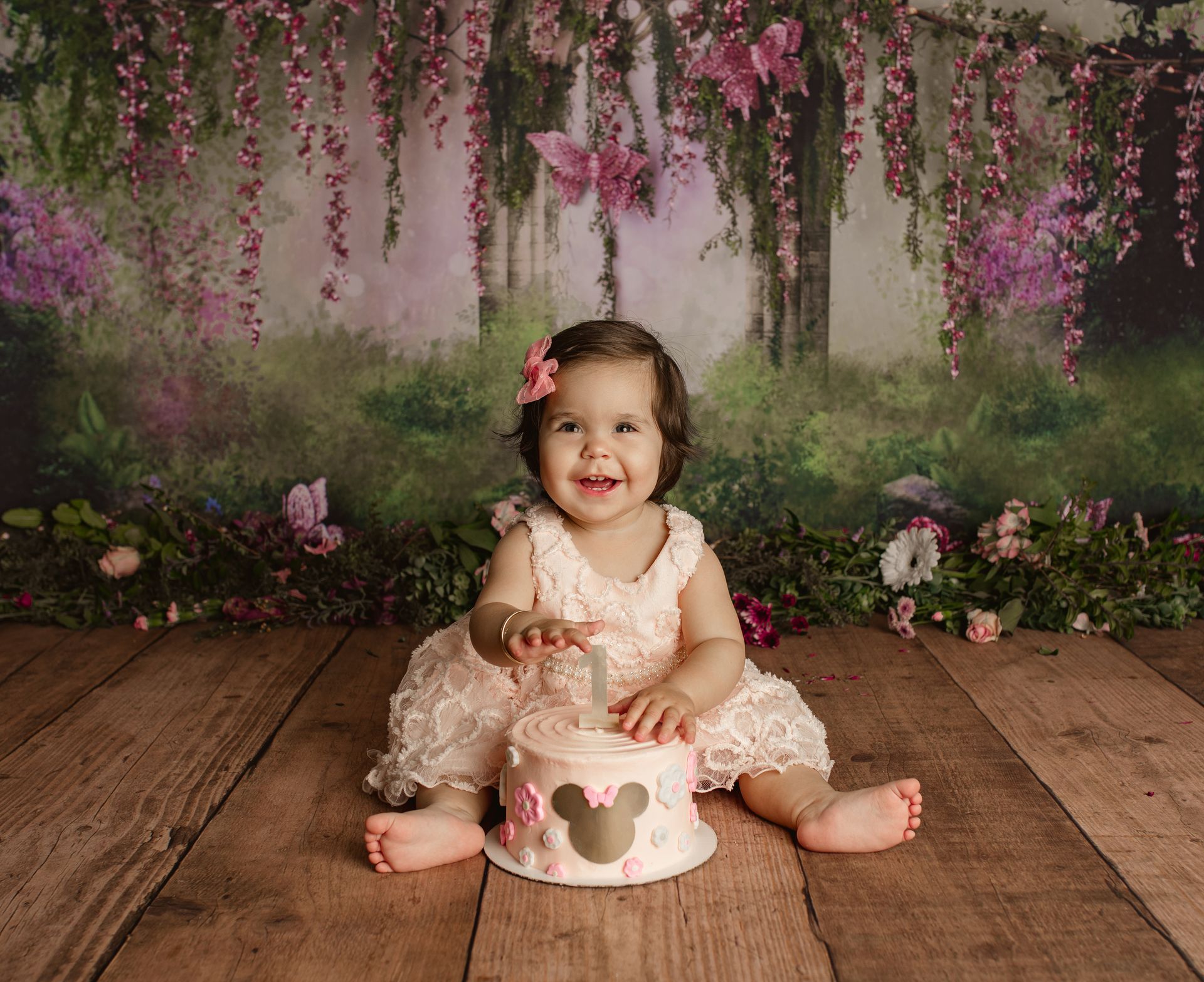 Smiling child in a cream dress sitting beside a heart-decorated cake on a wooden floor with flowers hanging above