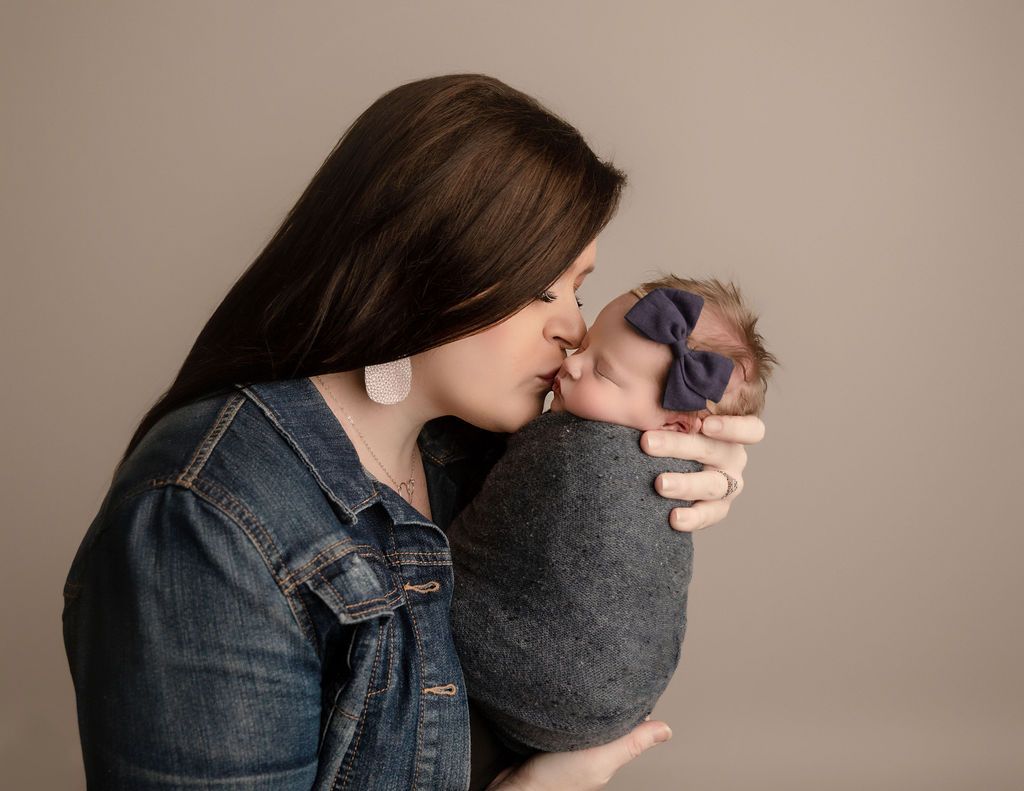 Woman in denim jacket kissing a swaddled baby with a blue bow. Gray background.