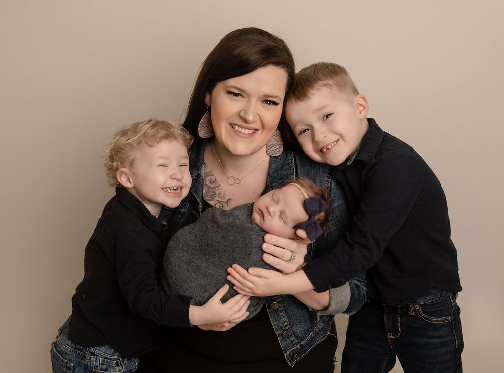 Woman in denim jacket hugs three children, one newborn, all smiling. Beige background.