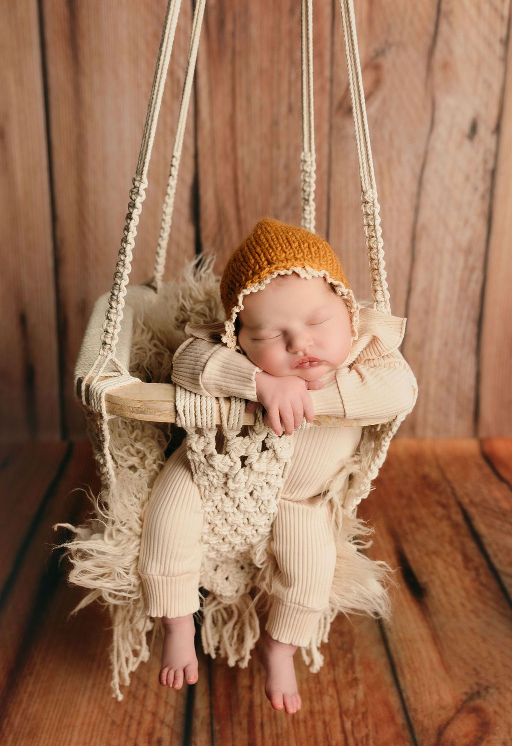 Newborn baby in crocheted hat and outfit, sleeping in a macrame swing against a wood background.