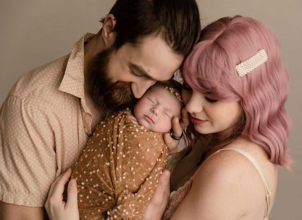 Parents cradling newborn wrapped in brown blanket; neutral background.