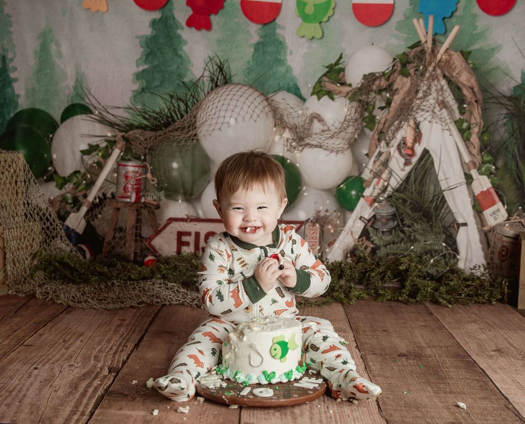 Boy in pajamas with cake, smiling. Rustic fishing-themed birthday setup with balloons, teepee, and wood backdrop.