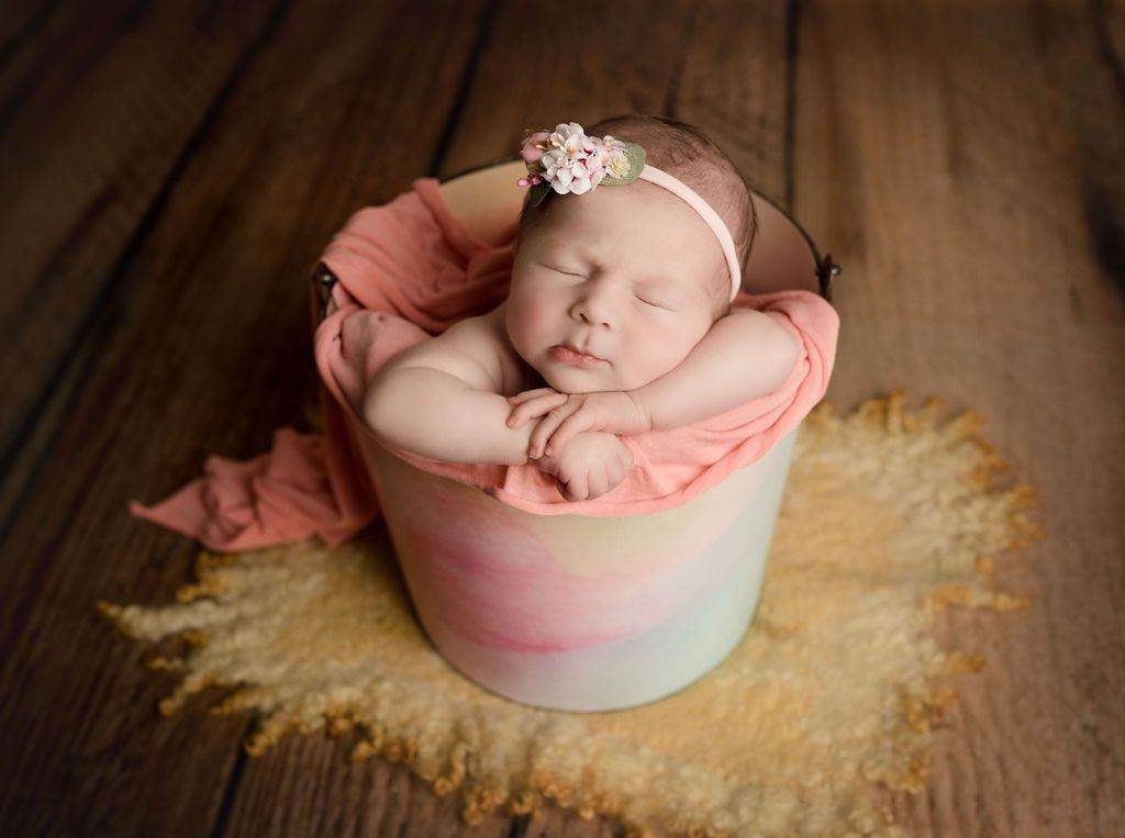 Newborn baby sleeping in a painted bucket, wearing a floral headband and draped with pink fabric, on a sheepskin rug.