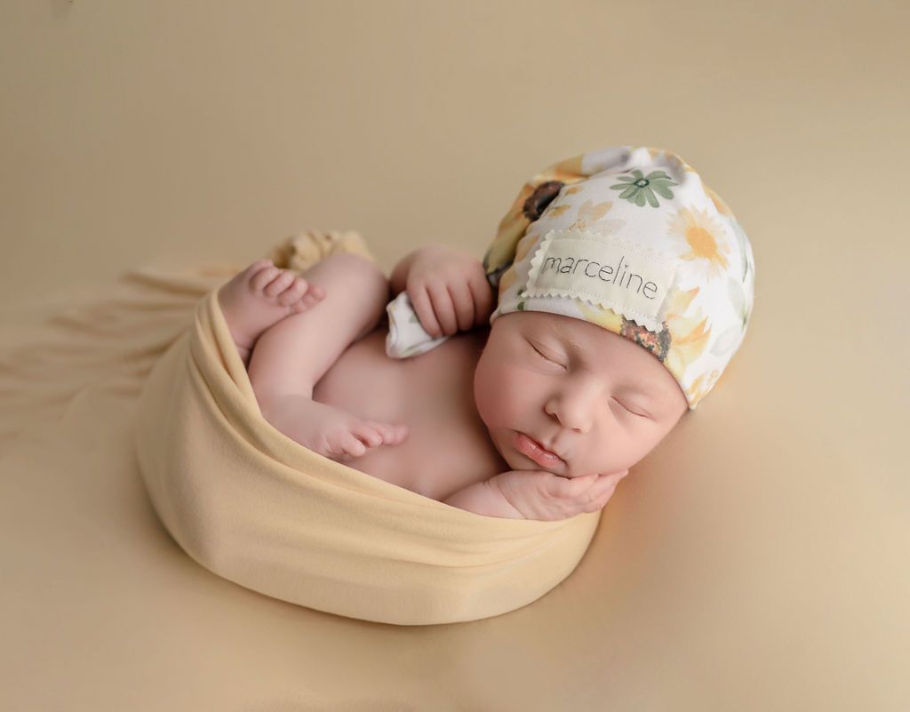 Newborn baby, swaddled in cream fabric, wearing a floral hat, lying on a beige surface.
