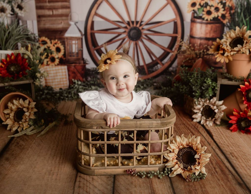 Baby in a wooden basket smiles, surrounded by sunflowers in a rustic setting with a large wooden wheel.