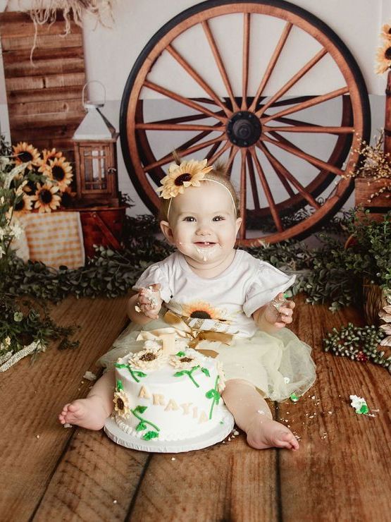 Baby with sunflower on head, smiling, covered in cake, in front of a wagon wheel.