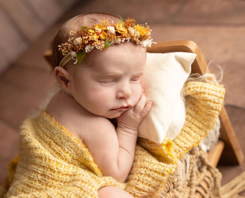 Newborn sleeps, wearing a floral crown, wrapped in a yellow blanket, resting on a pillow.