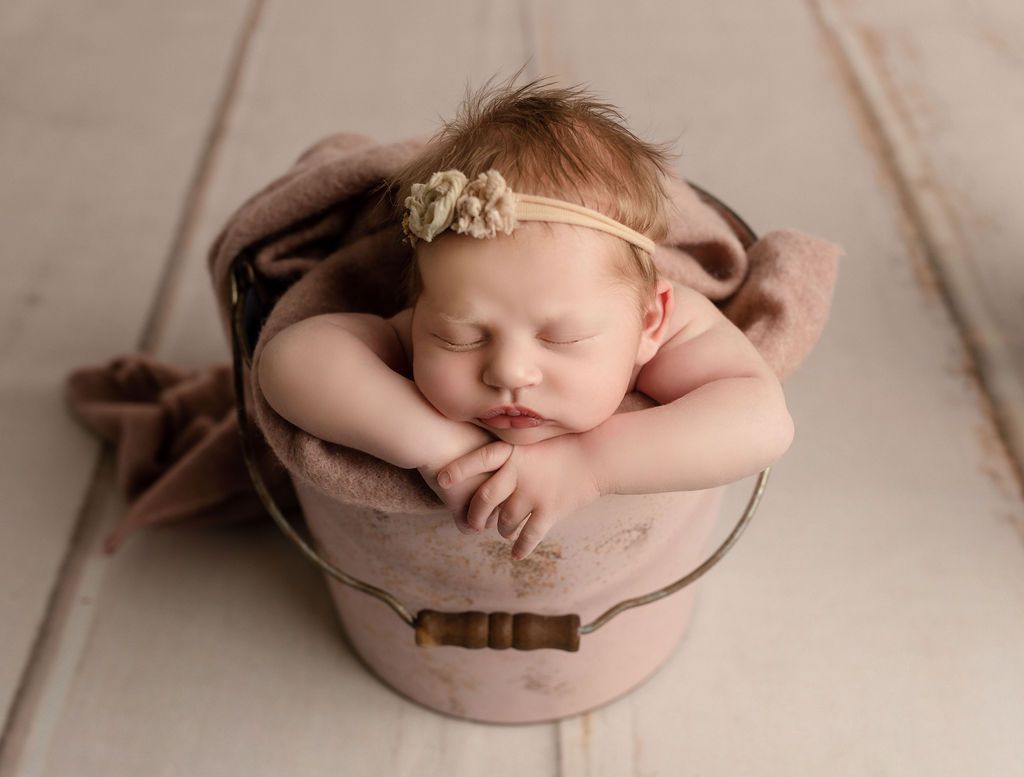 Newborn baby sleeping in a pink bucket, wrapped in a blanket, wearing a headband.