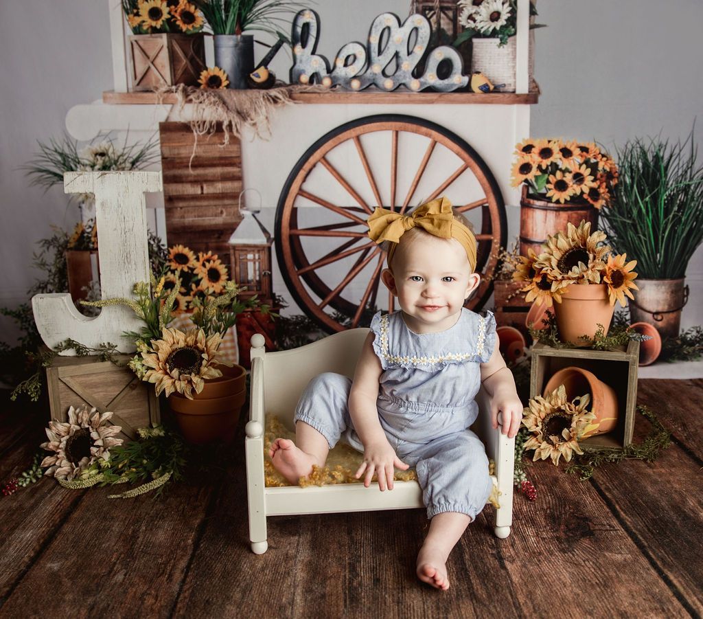 Baby in blue jumpsuit and yellow bow sits on miniature bed in sunflower-themed setting.