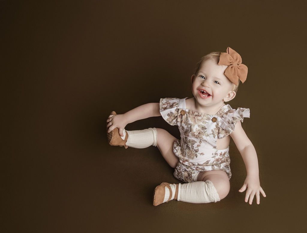 Smiling toddler in floral romper and bow, seated on brown surface, reaching for foot.