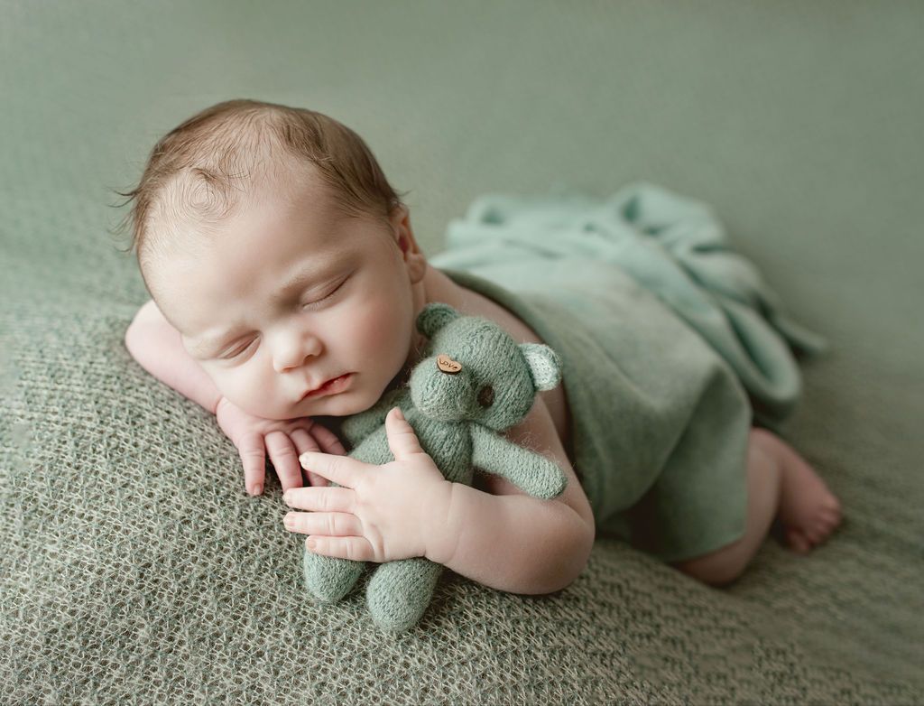 Newborn sleeping, wrapped in green, cuddles a green teddy bear on a green blanket.