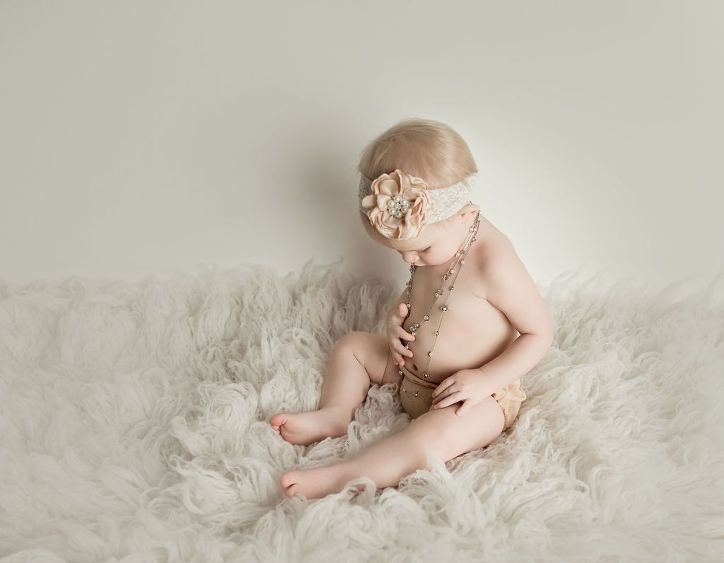 Baby sitting on white, fluffy material, wearing a headband and beaded necklace, looking down.