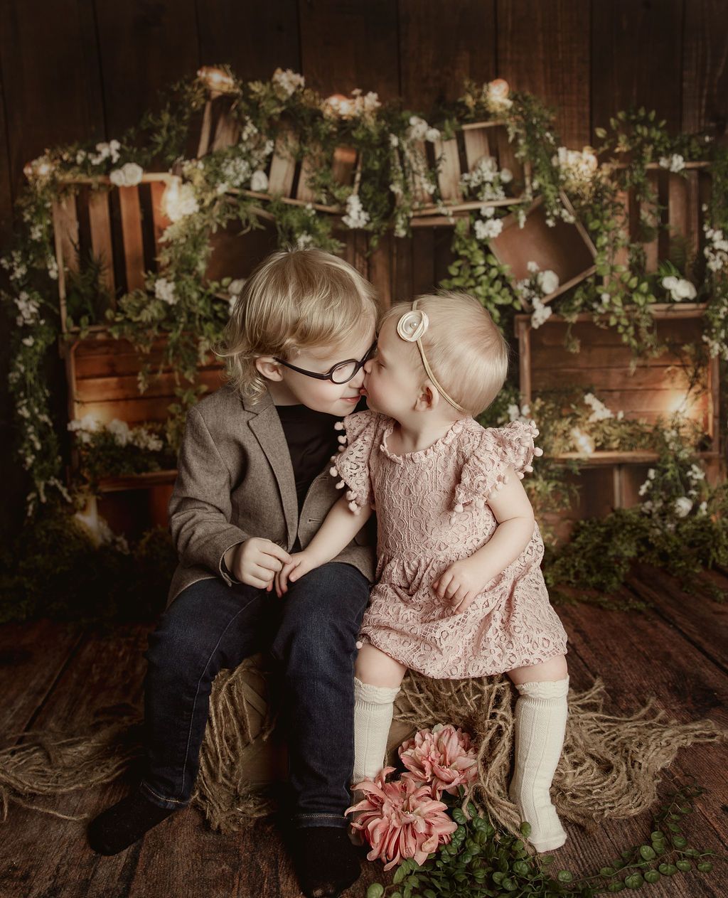 Boy with glasses and a girl in a dress touch noses, seated on straw. Floral backdrop with crates.
