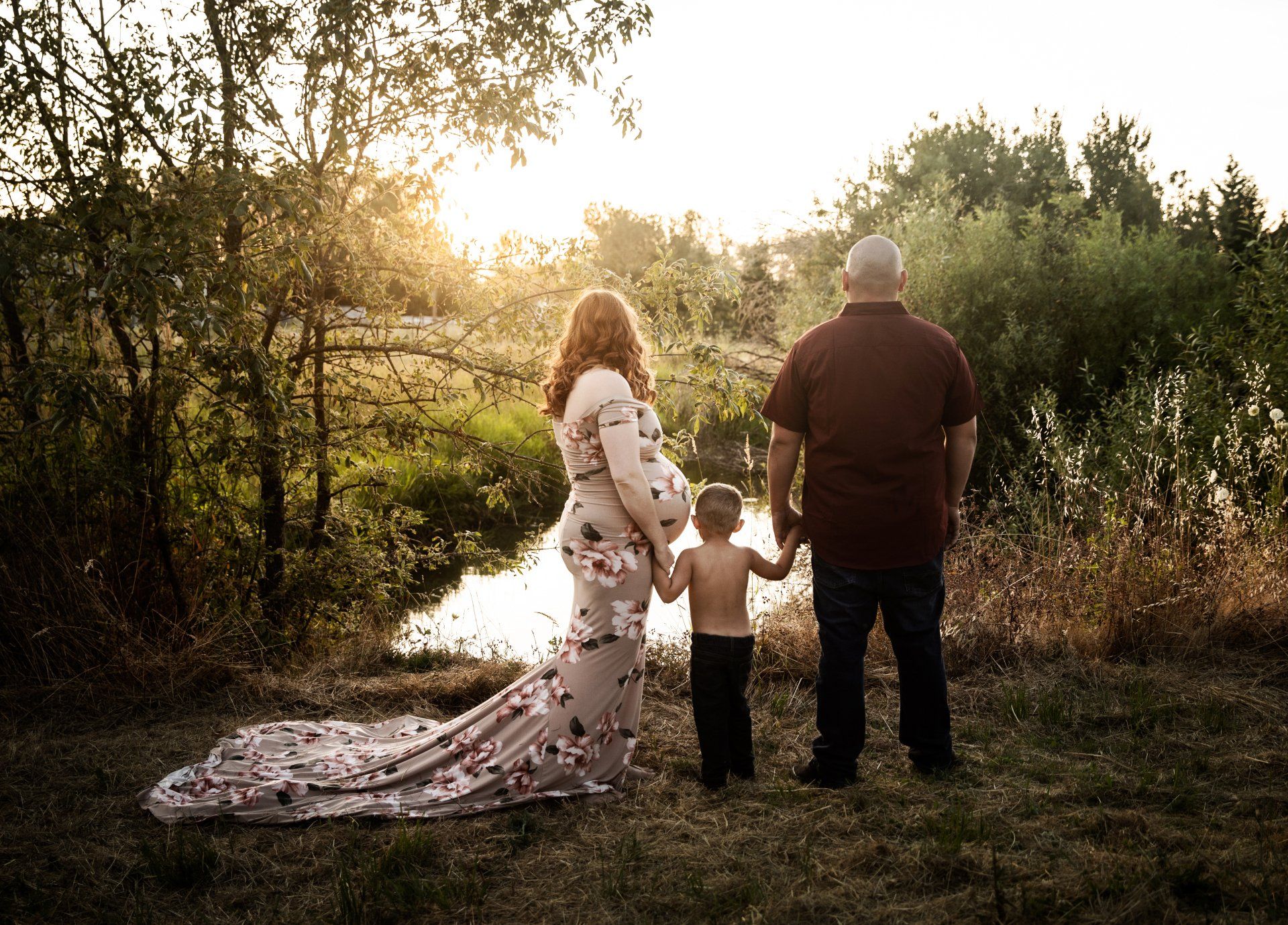 Pregnant person, child, and adult standing by a stream at sunset; the family holds hands.