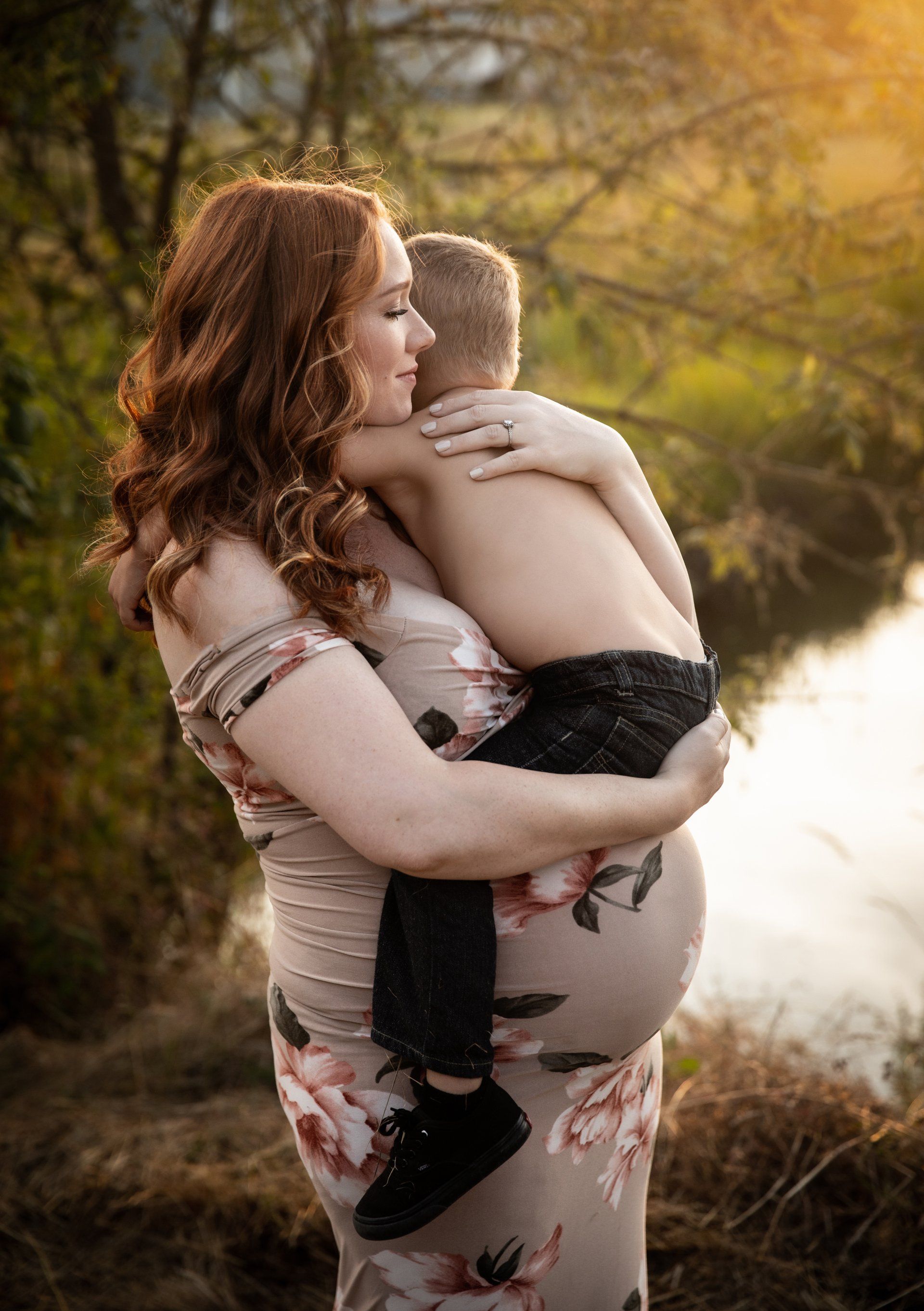 Pregnant person with reddish-brown hair hugs a child near a body of water; they are outdoors.