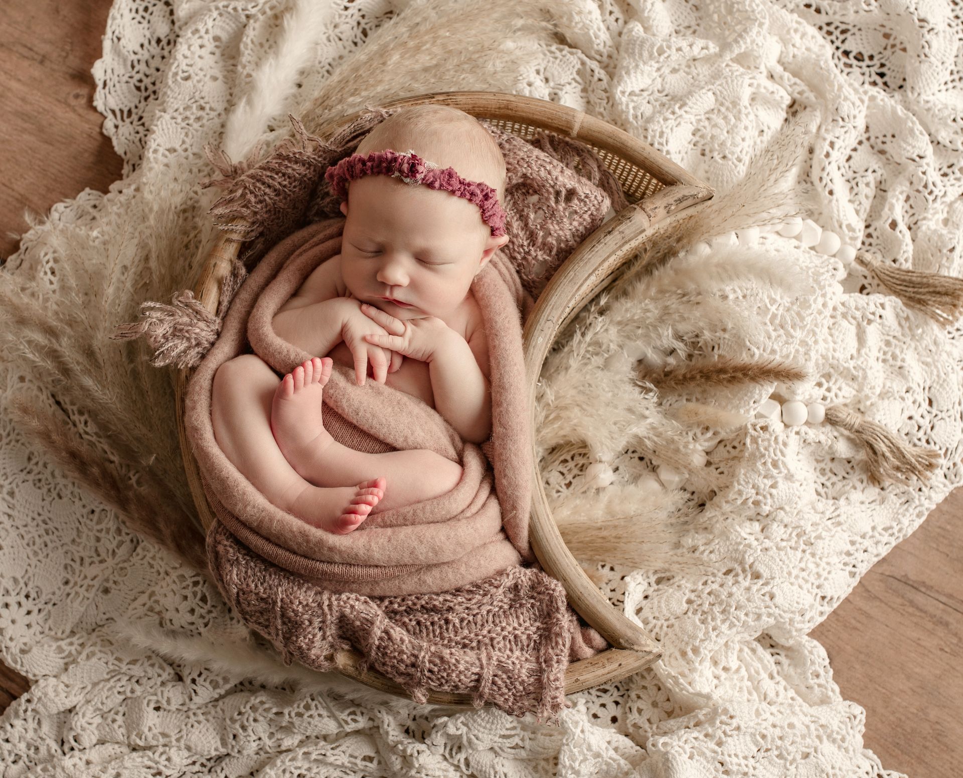 Newborn baby swaddled in pink blanket, wearing floral headband, nestled in woven basket with dried plants.