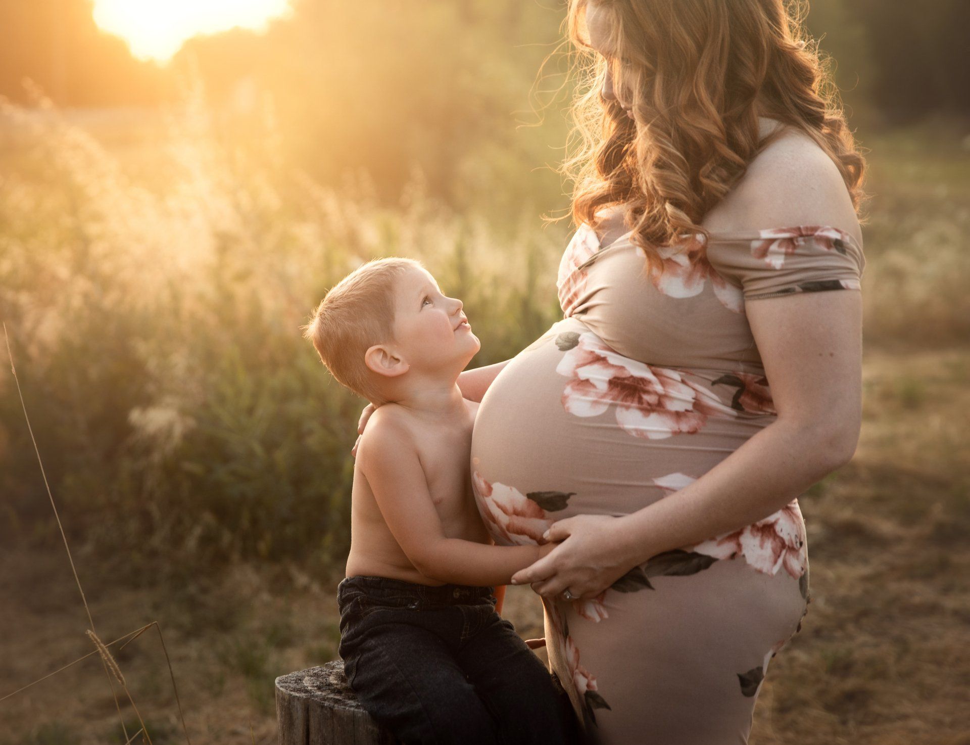 Pregnant woman in floral dress, young child looking up and touching her belly in a sunlit field.