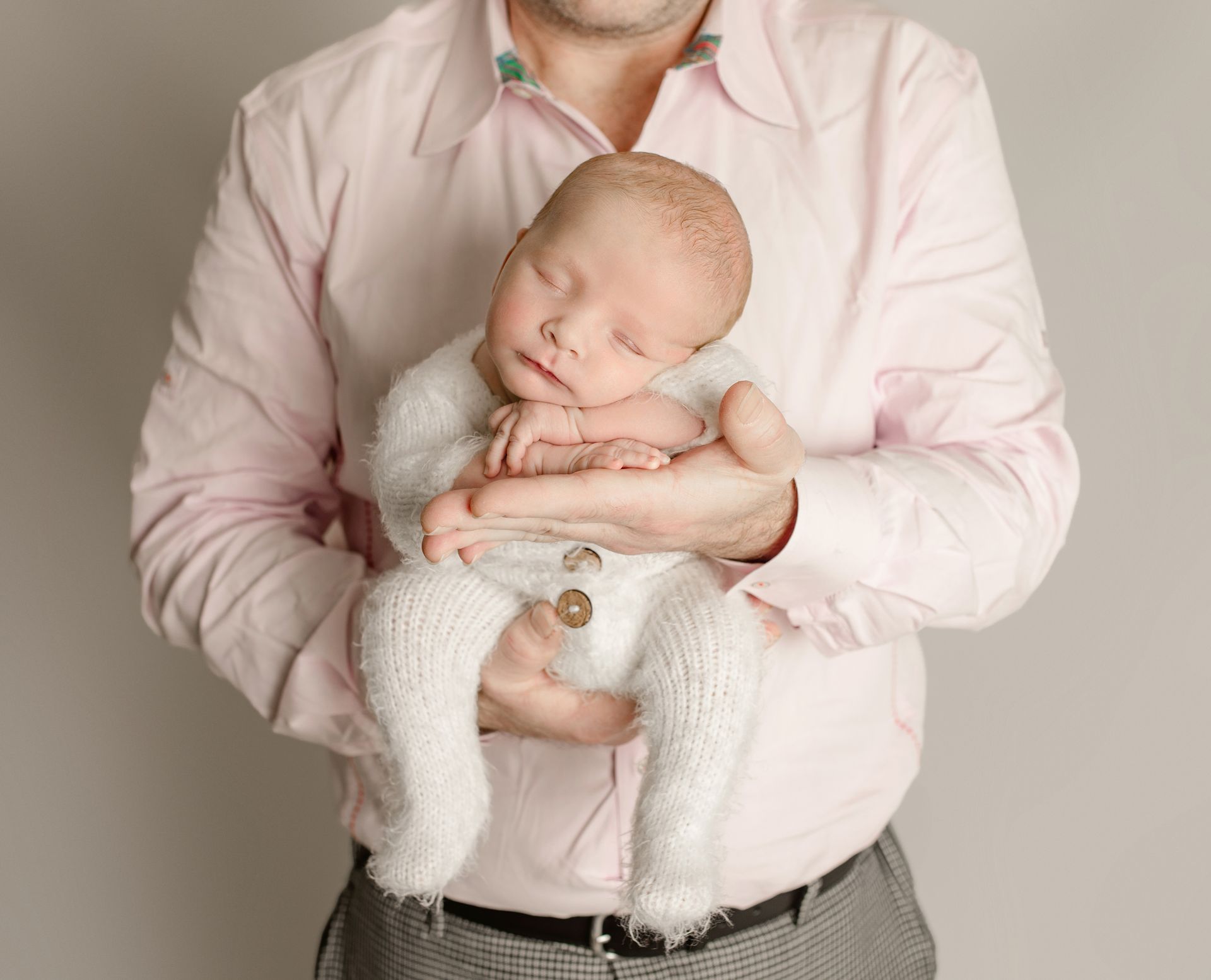 A baby wearing white knit outfit is held by a person in a pink shirt.