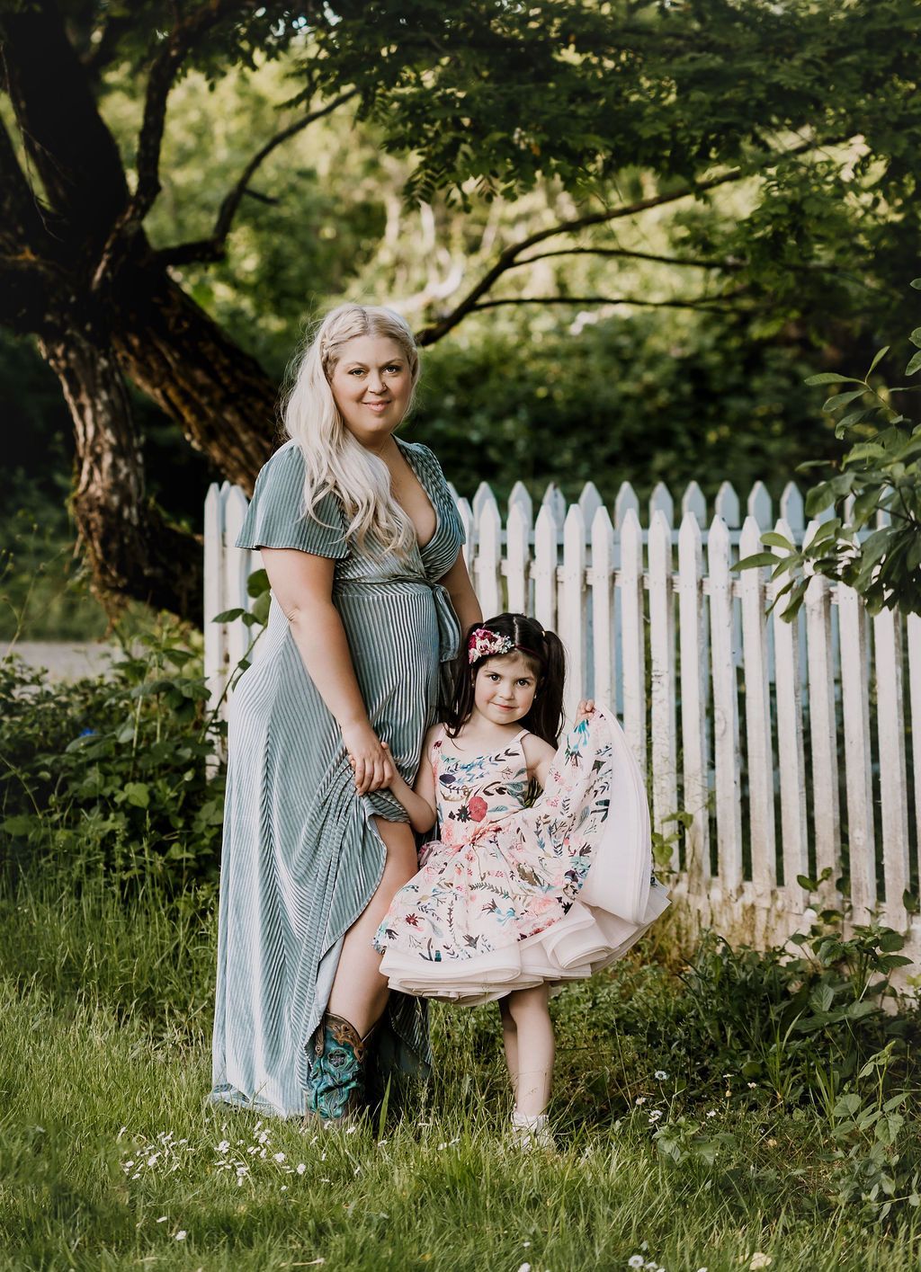 Woman and child holding hands in front of a white picket fence, woman in blue striped dress.