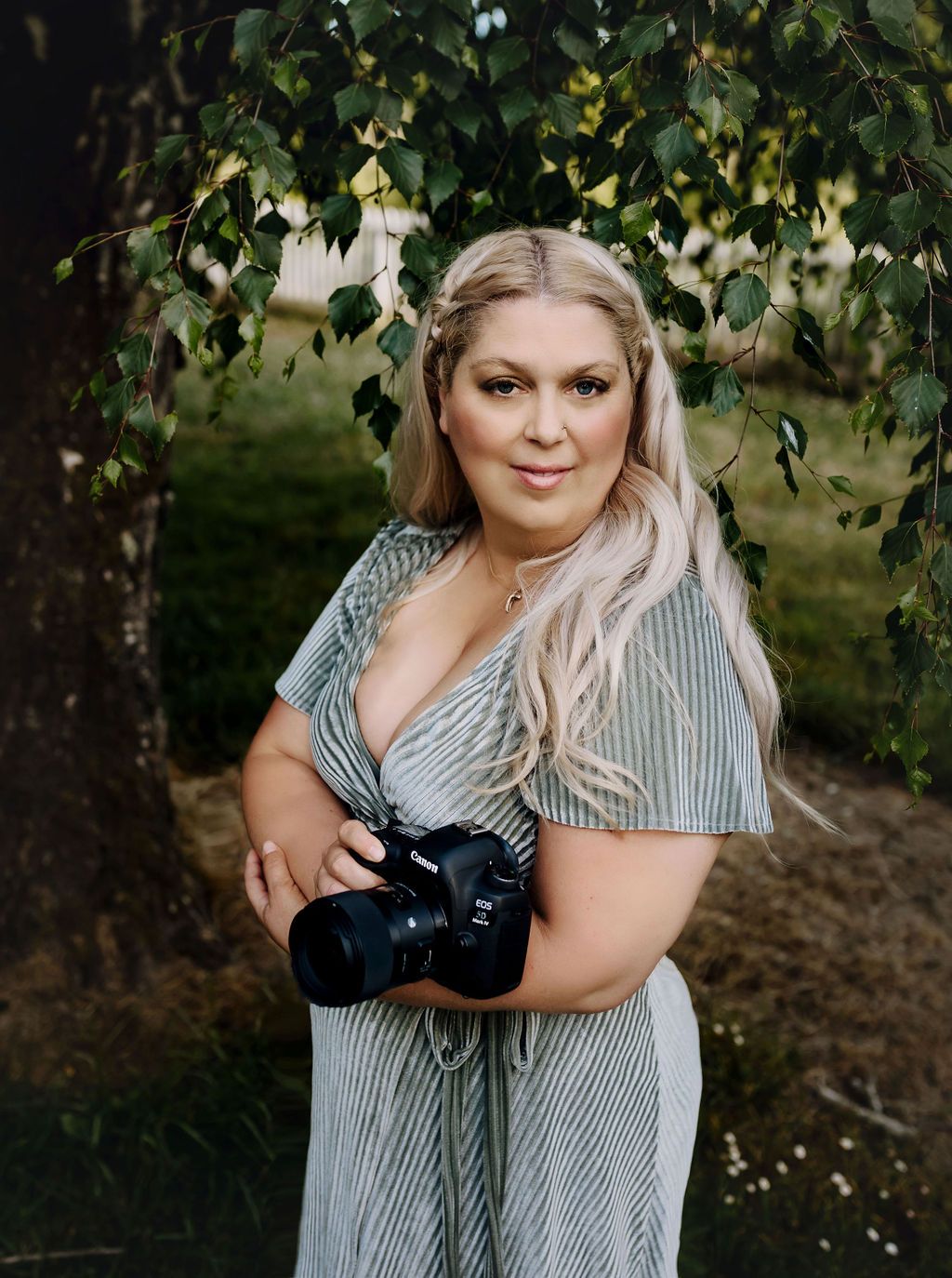 Woman in green dress holding camera, standing near a tree in a natural setting.