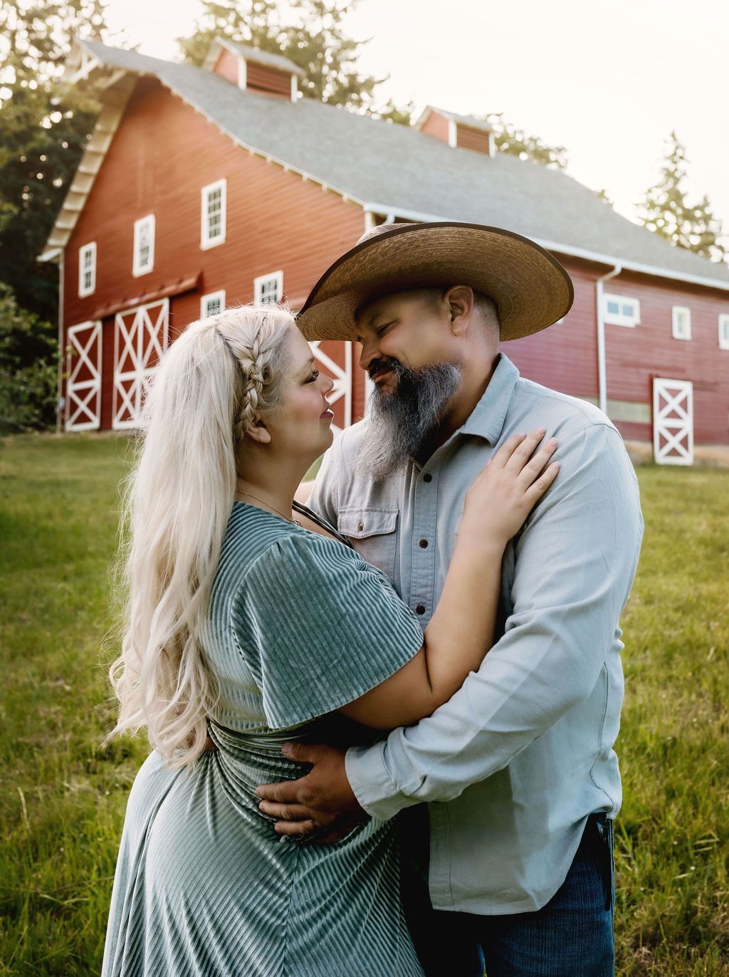 Couple embracing in front of a red barn, looking at each other. Woman has blonde hair and a green dress, man wears a hat.