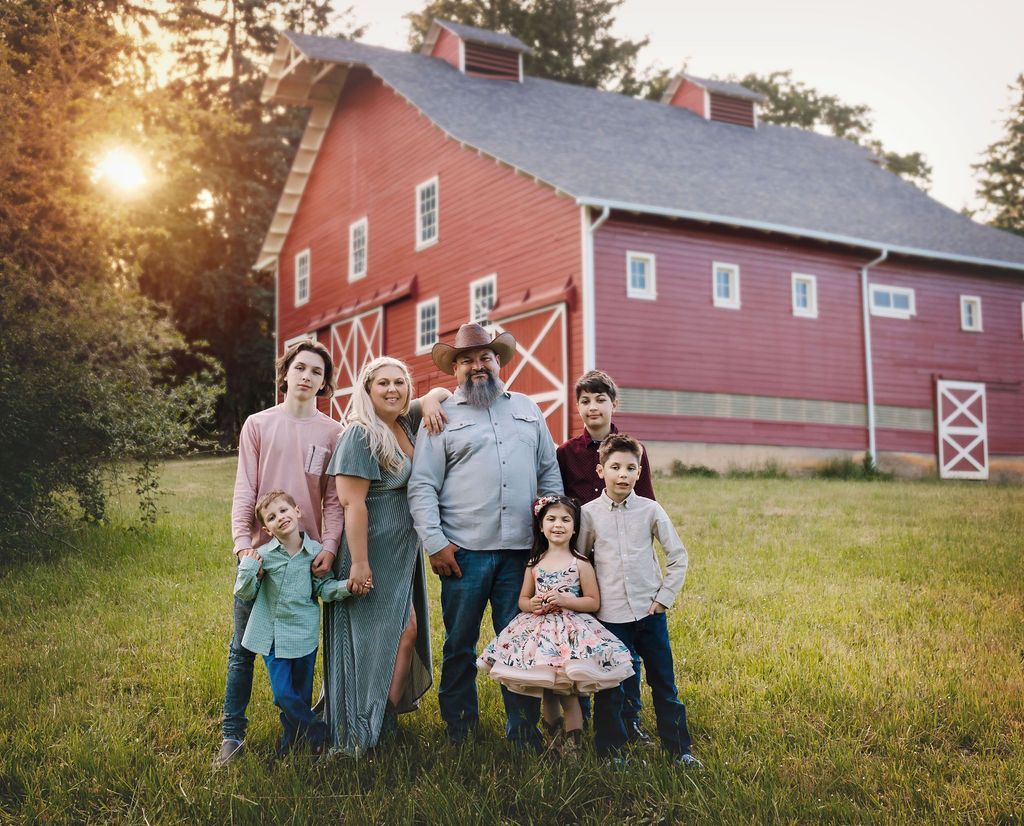 Family of seven smiling in front of a red barn with golden sunlight.