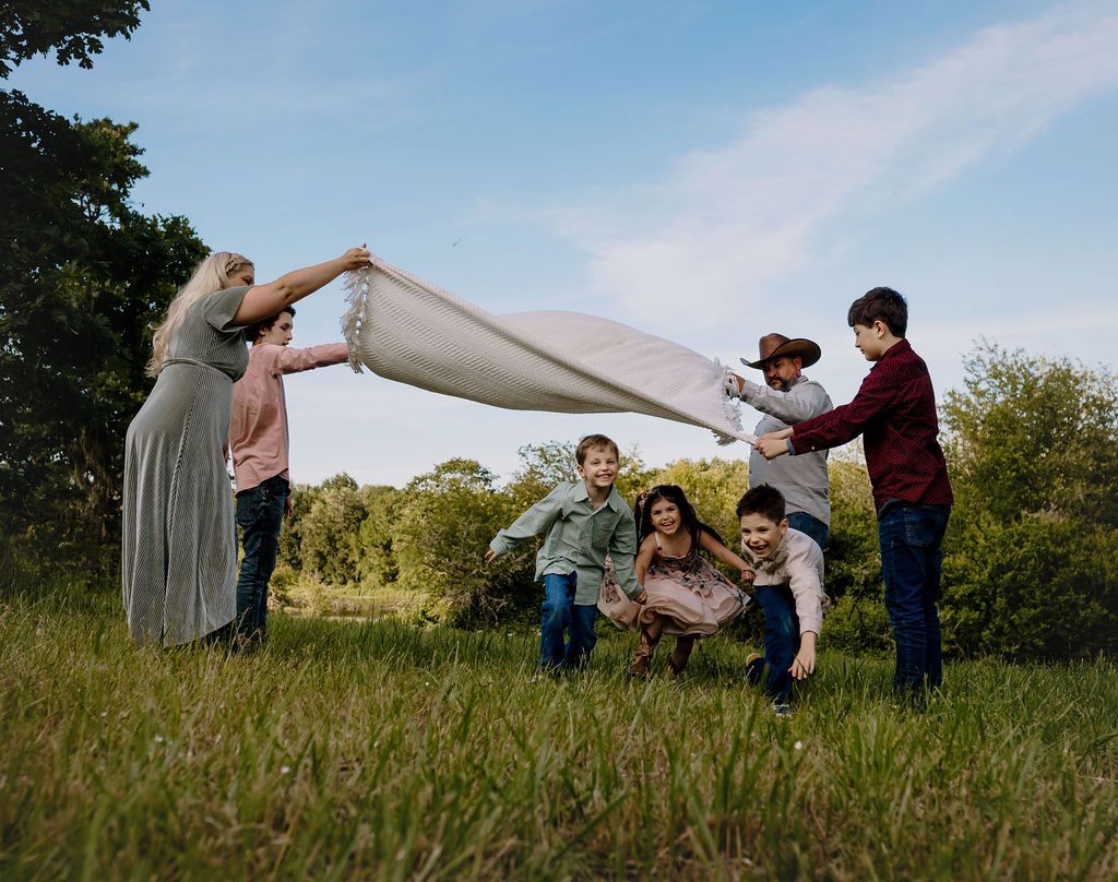 Family outdoors with blanket, holding it up while kids run toward it in the grass.