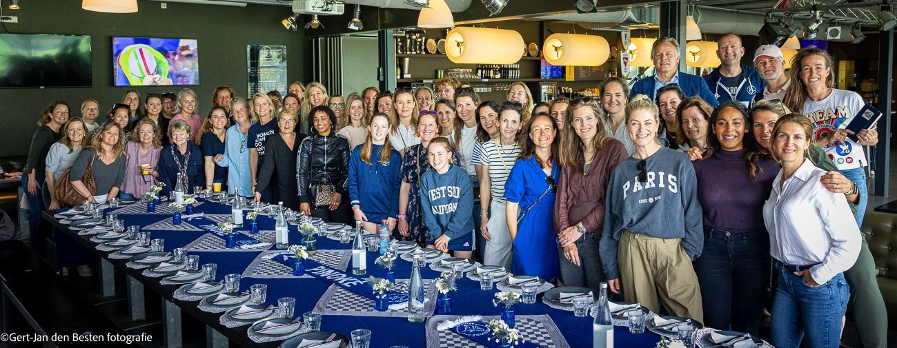 Een grote groep poseert in een feestzaal naast lange tafels gedekt met blauwe tafelkleden en gevouwen servetten.