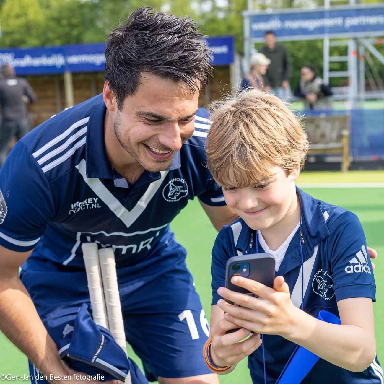 Een coach en een kind in blauwe shirts staan lachend naar een telefoon te kijken op een sportveld.