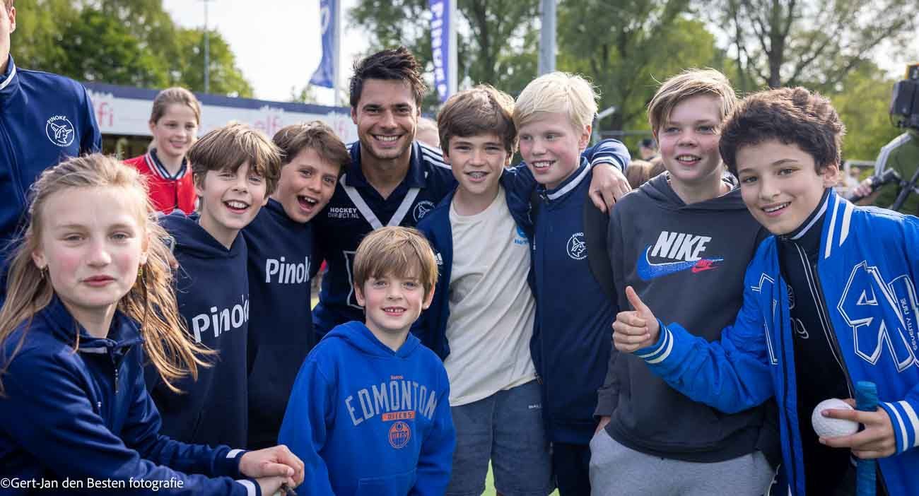 Een groep lachende mensen in blauwe sporttruien poseert buiten voor bomen.