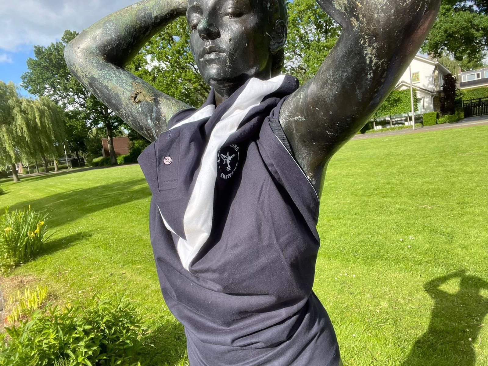 Bronzen beeld met opgeheven armen in een groen park, gekleed in een donker shirt en een witte sjaal.