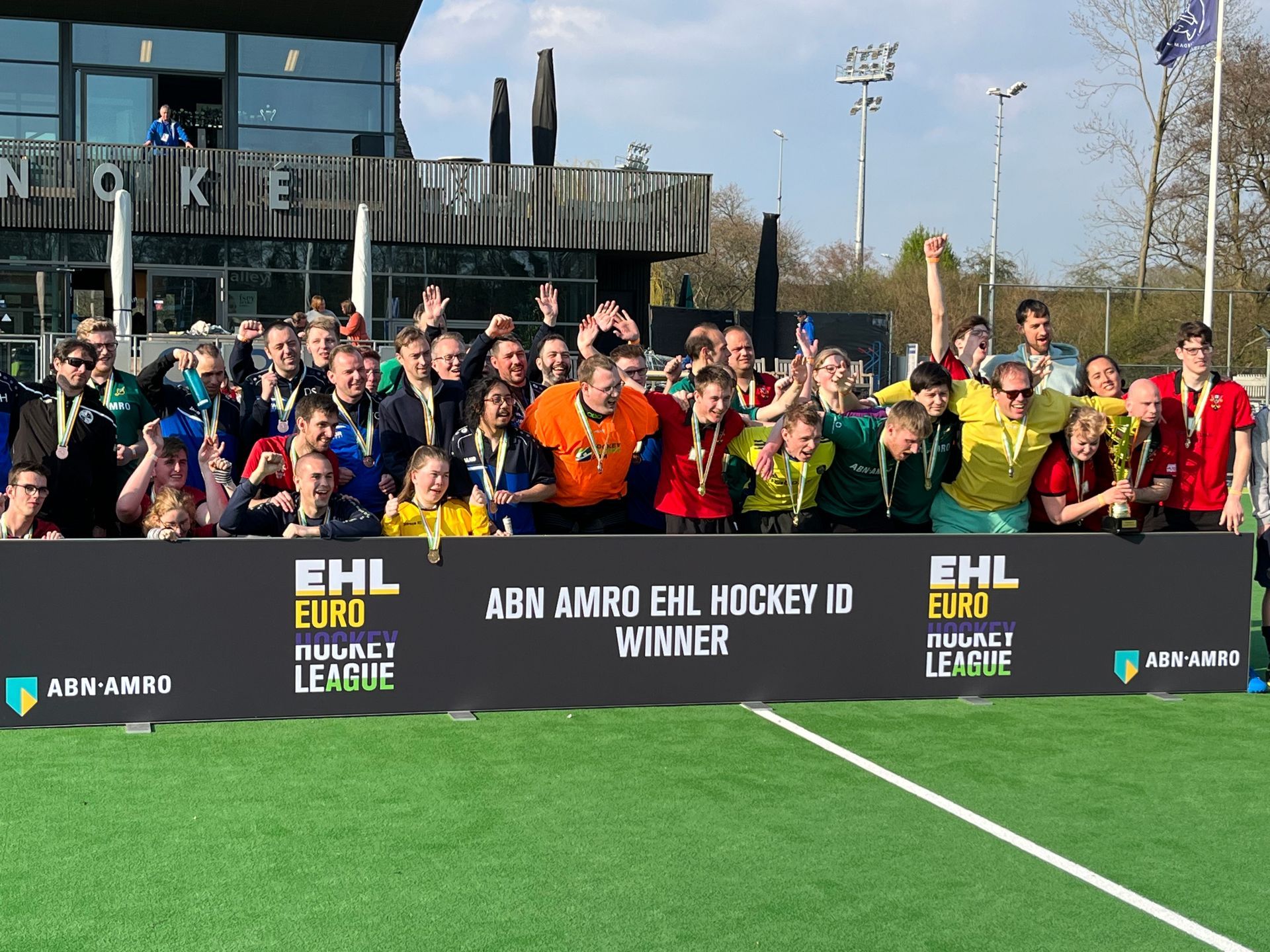 Een groep mensen poseert achter een spandoek van de EHL Field Hockey League op een kunstgrasveld in de buitenlucht.