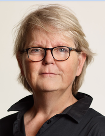 Portret van een persoon met kort blond haar, een zwarte bril en een donker overhemd met kraag, op een effen achtergrond.