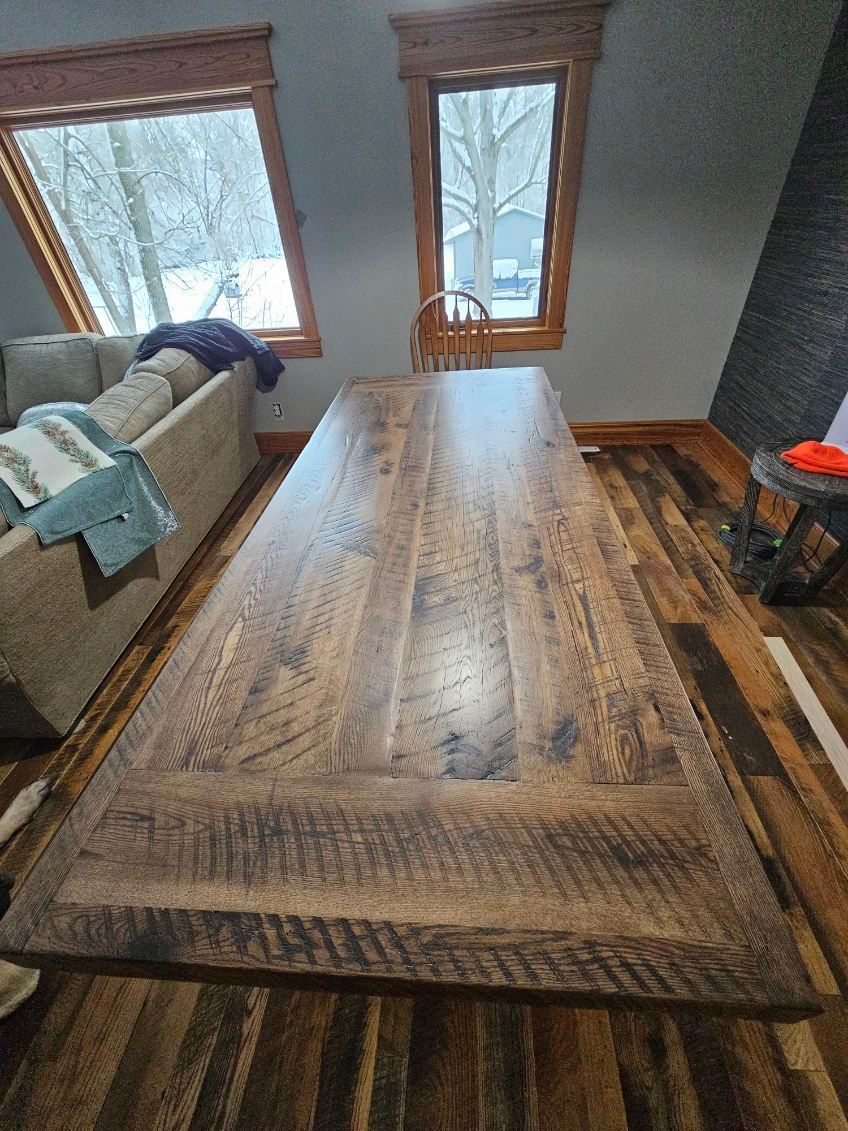 Large, weathered wooden dining table in a room with hardwood floors and windows overlooking a snowy landscape.
