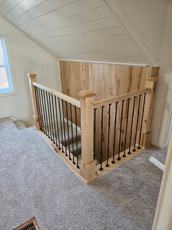 Staircase with light wood railing and black spindles, carpeted floor, and wooden wall paneling in a room with angled ceiling.