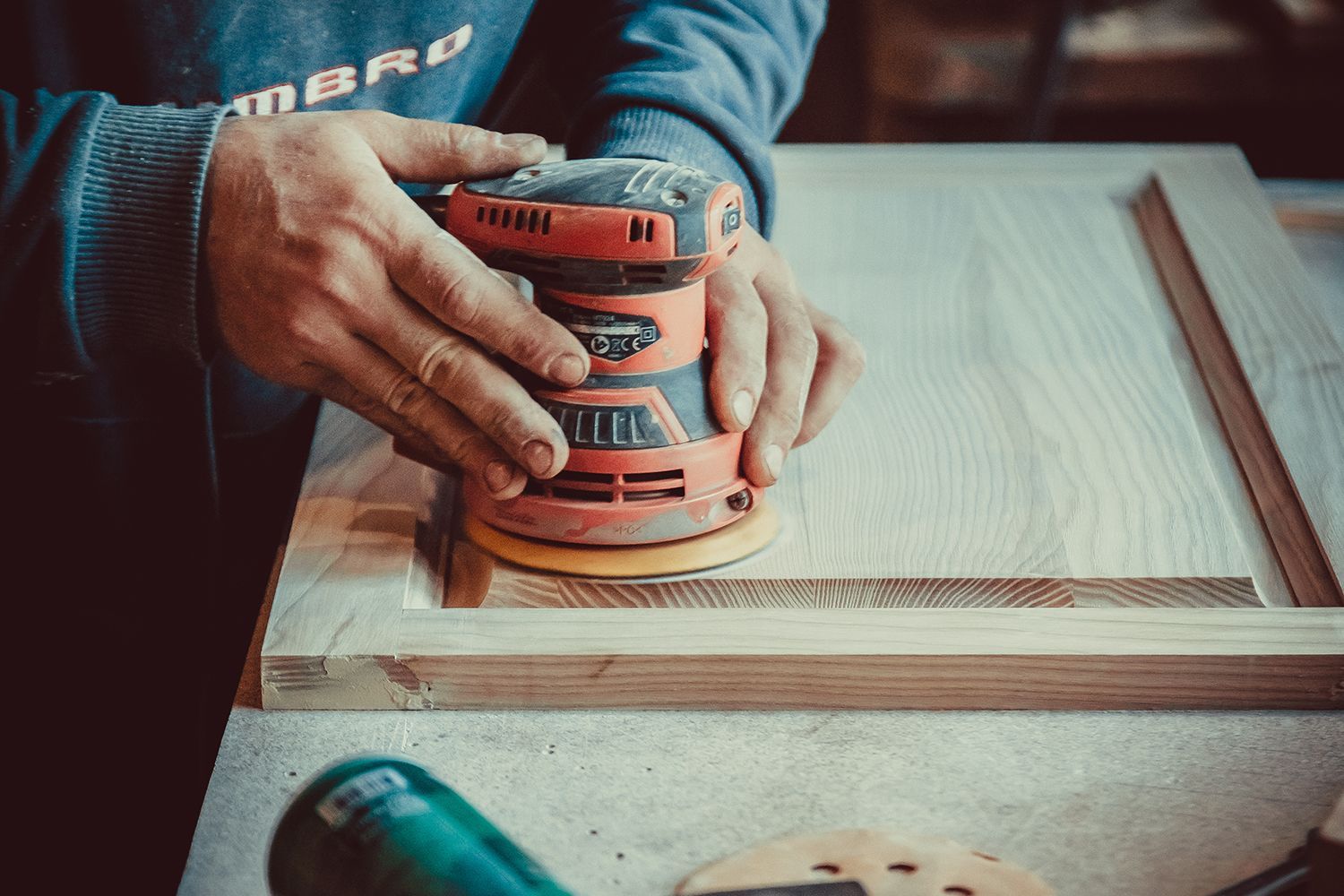 Person sanding wooden door with an orbital sander in a workshop.