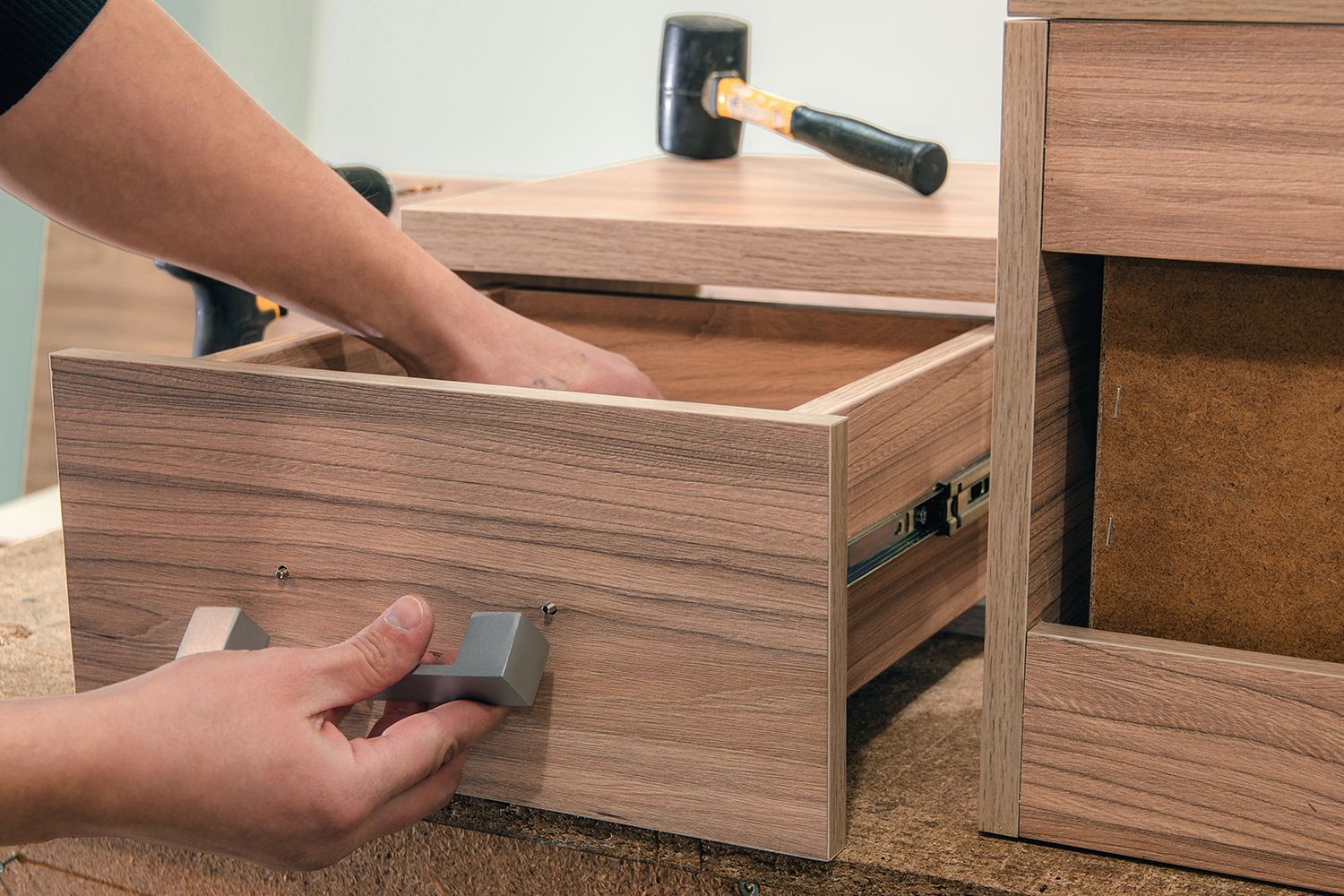Person assembling a wooden drawer, with a hammer in the background.