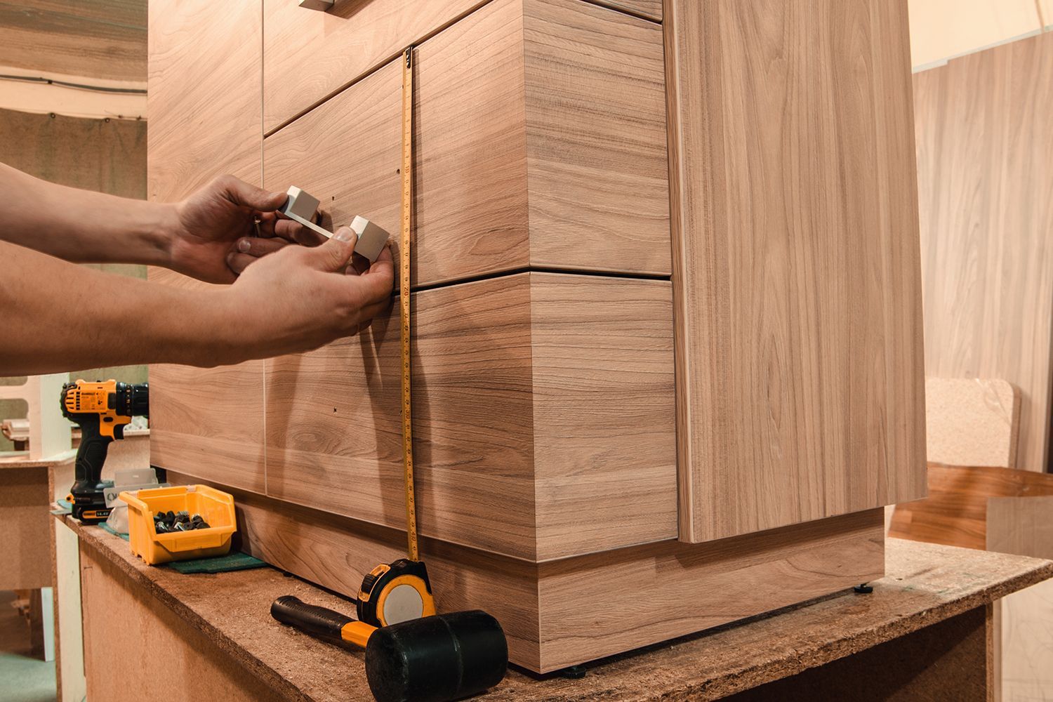 Person installing drawer pulls on a wooden cabinet; tools and tape measure visible.