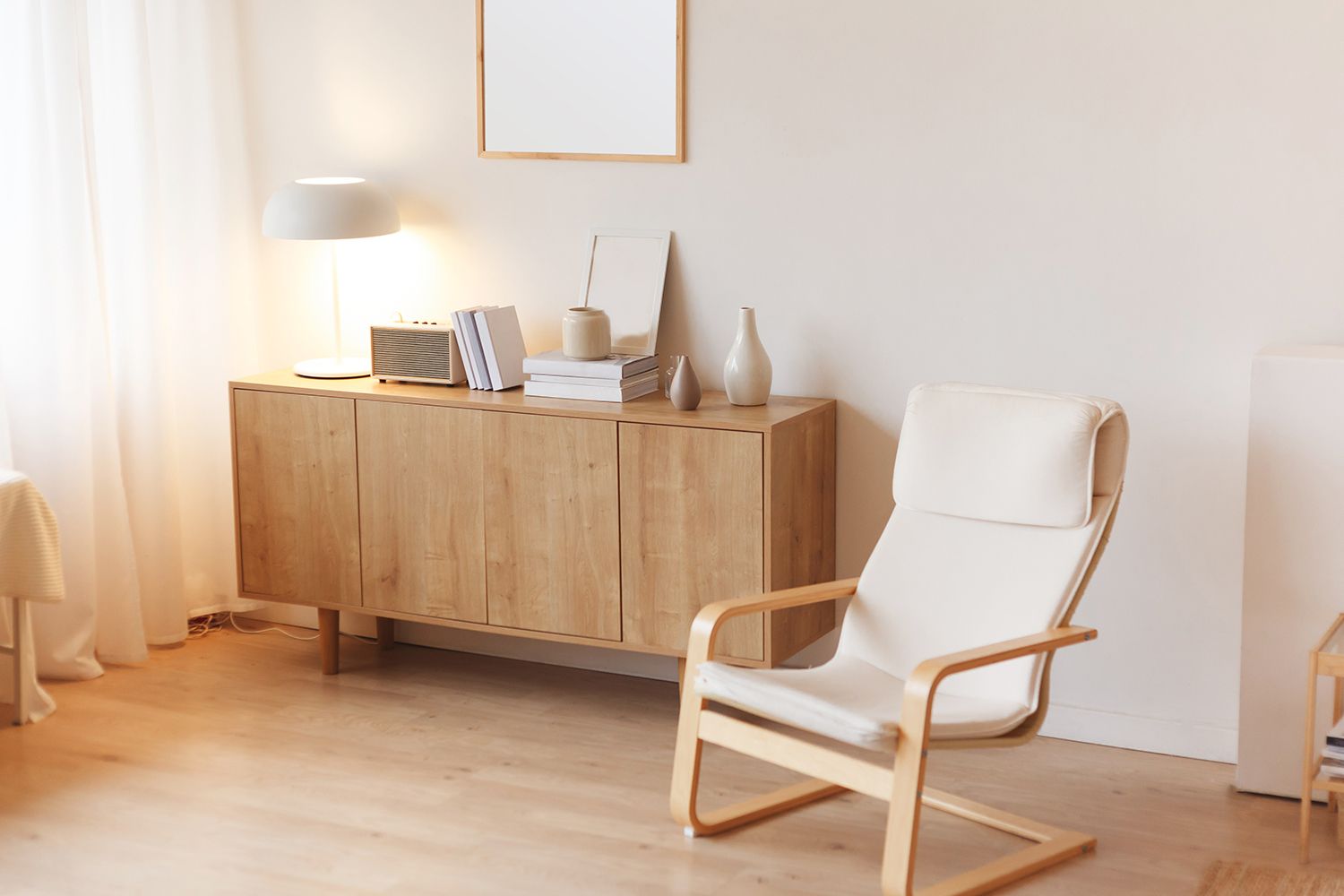 Wooden cabinet with lamp, books, and decor, beside a white armchair in a bright room.
