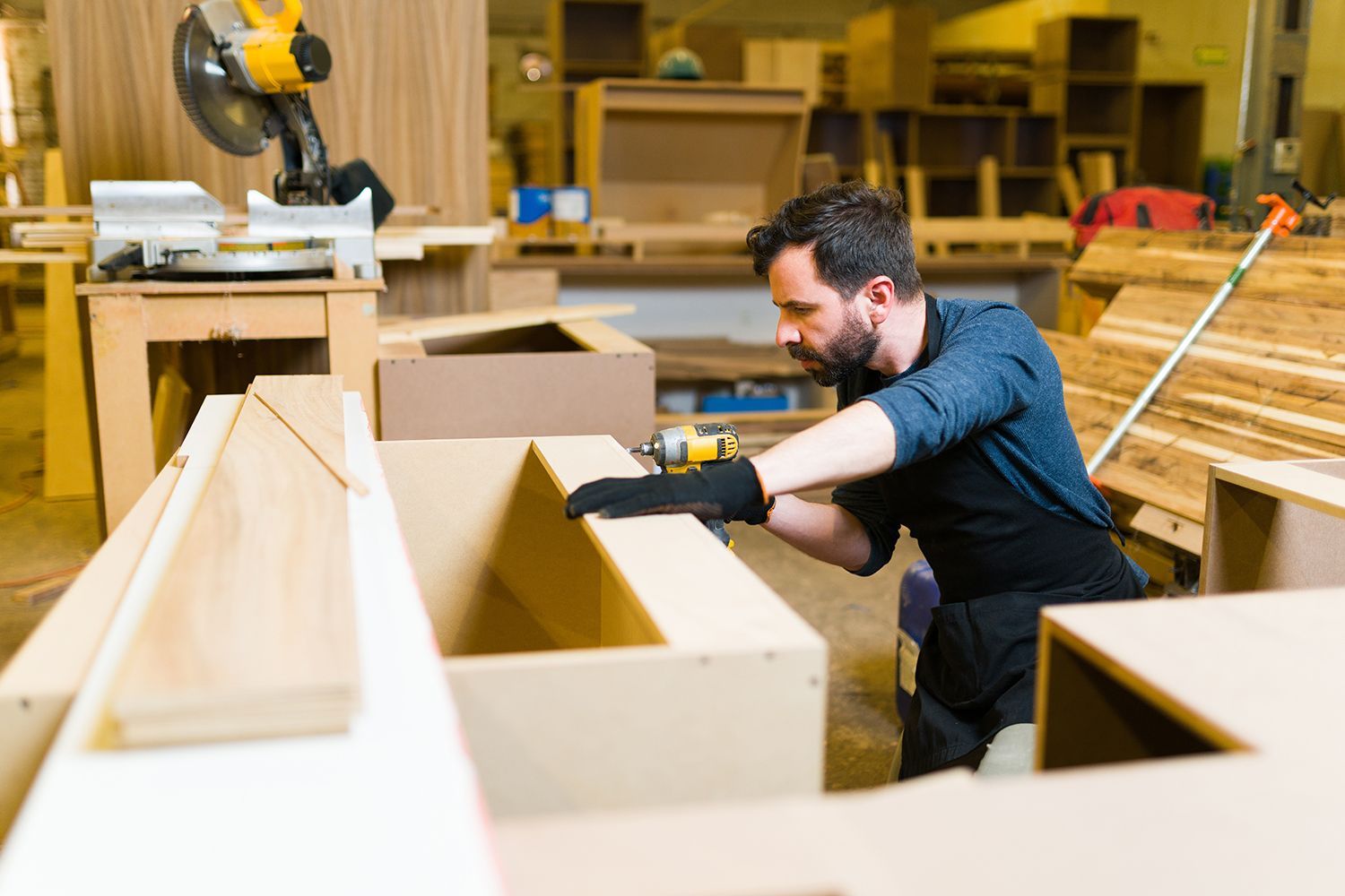 Man in workshop assembling wooden furniture with a power drill.