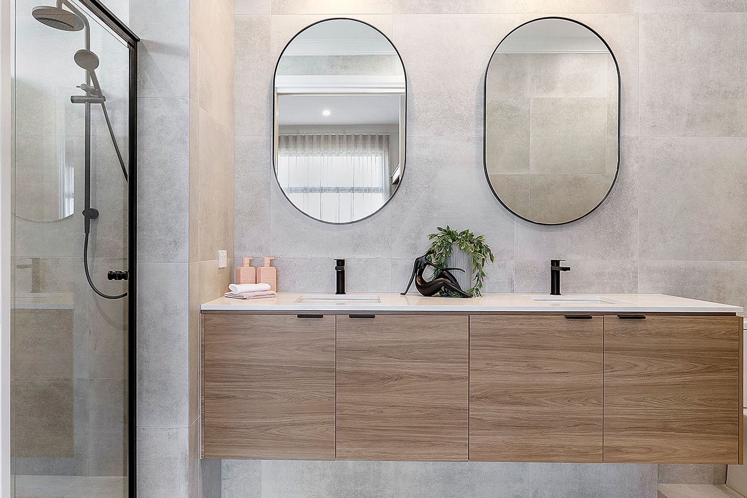 Modern bathroom with light wood vanity, two oval mirrors, black fixtures, and a shower.