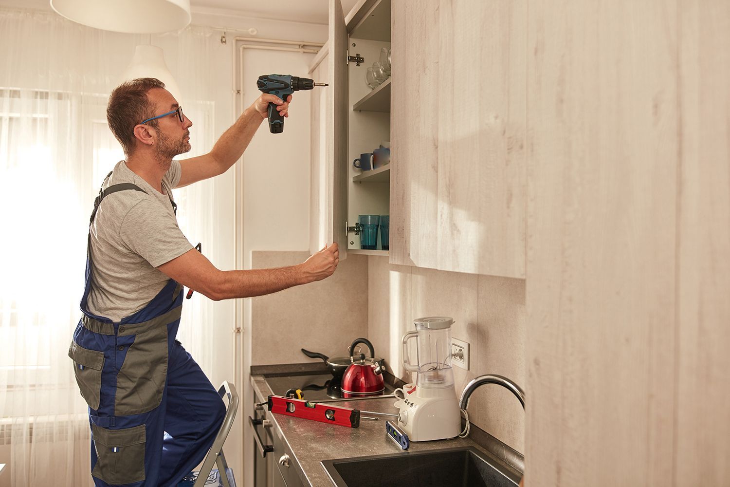 Man using a drill on a kitchen cabinet, standing on a step stool.