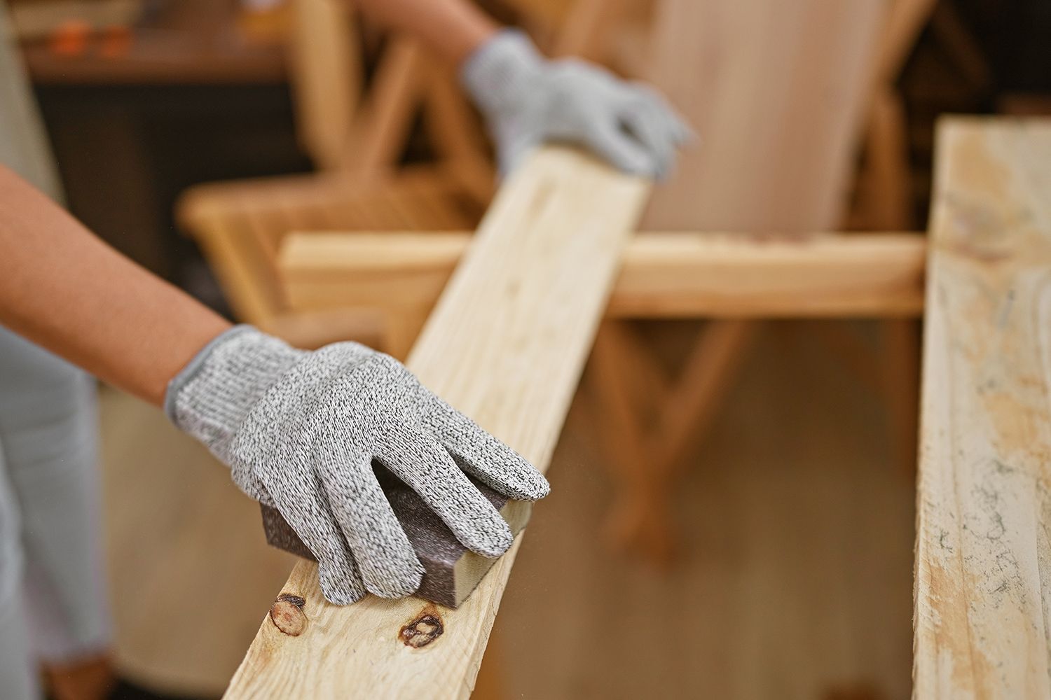 Person sanding a wooden plank with a sanding block, wearing gray gloves, in a workshop. Person sanding a wooden plank with a sanding block, wearing gray gloves, in a workshop.