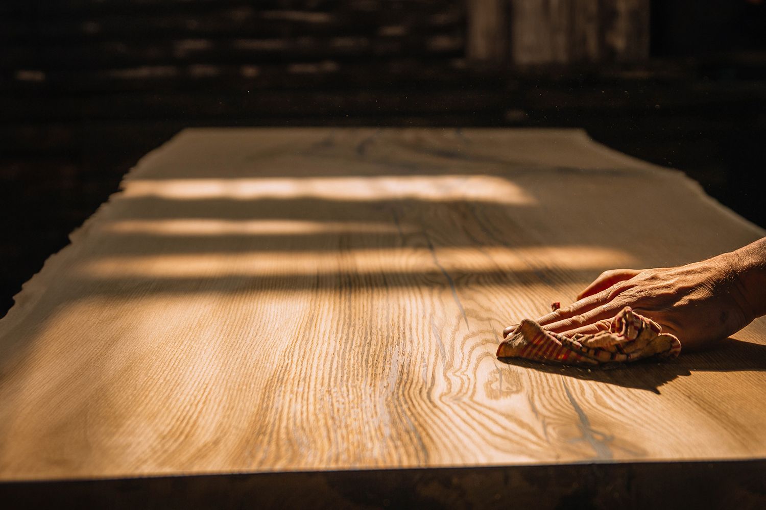 A hand sanding a large, unfinished wooden table, illuminated by sunlight.