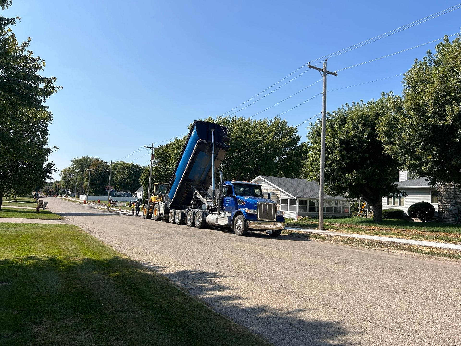 A blue semi-truck dumping gravel on a residential street; construction in progress.
