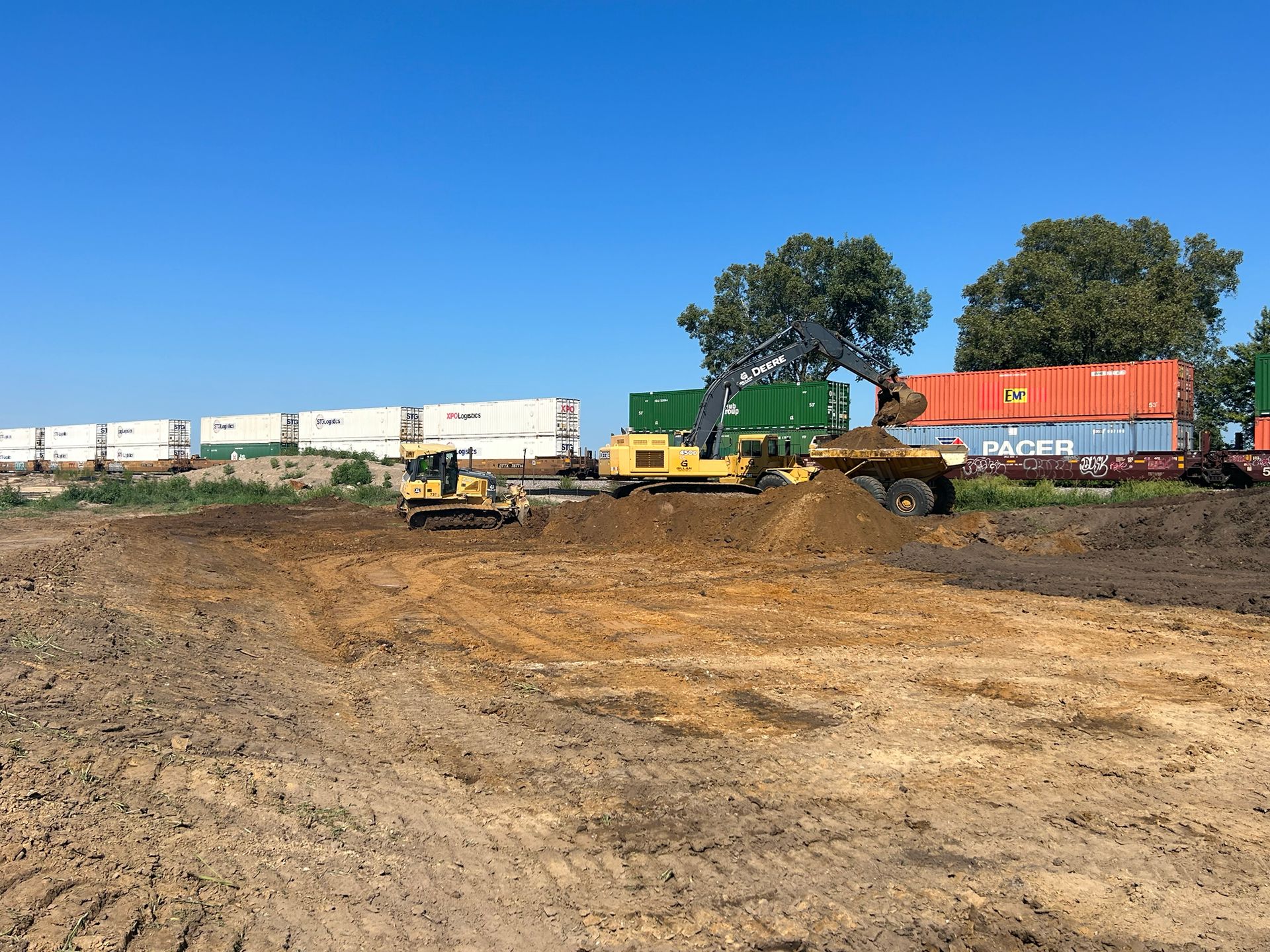 Construction site with bulldozer, excavator, and dump truck moving dirt; cargo containers in the background.