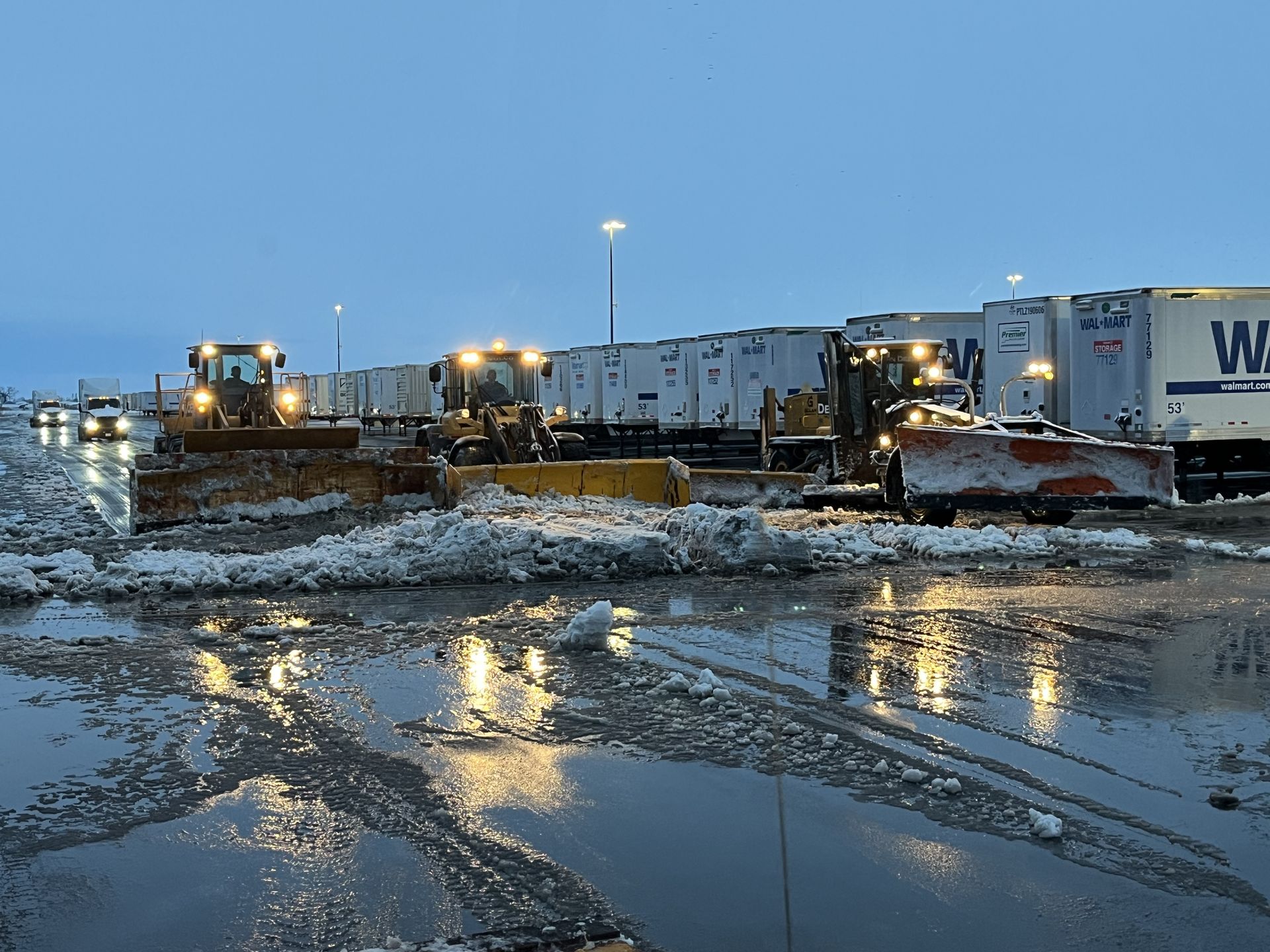 Snow plows clearing snow from a lot. Trucks are parked behind the plows.