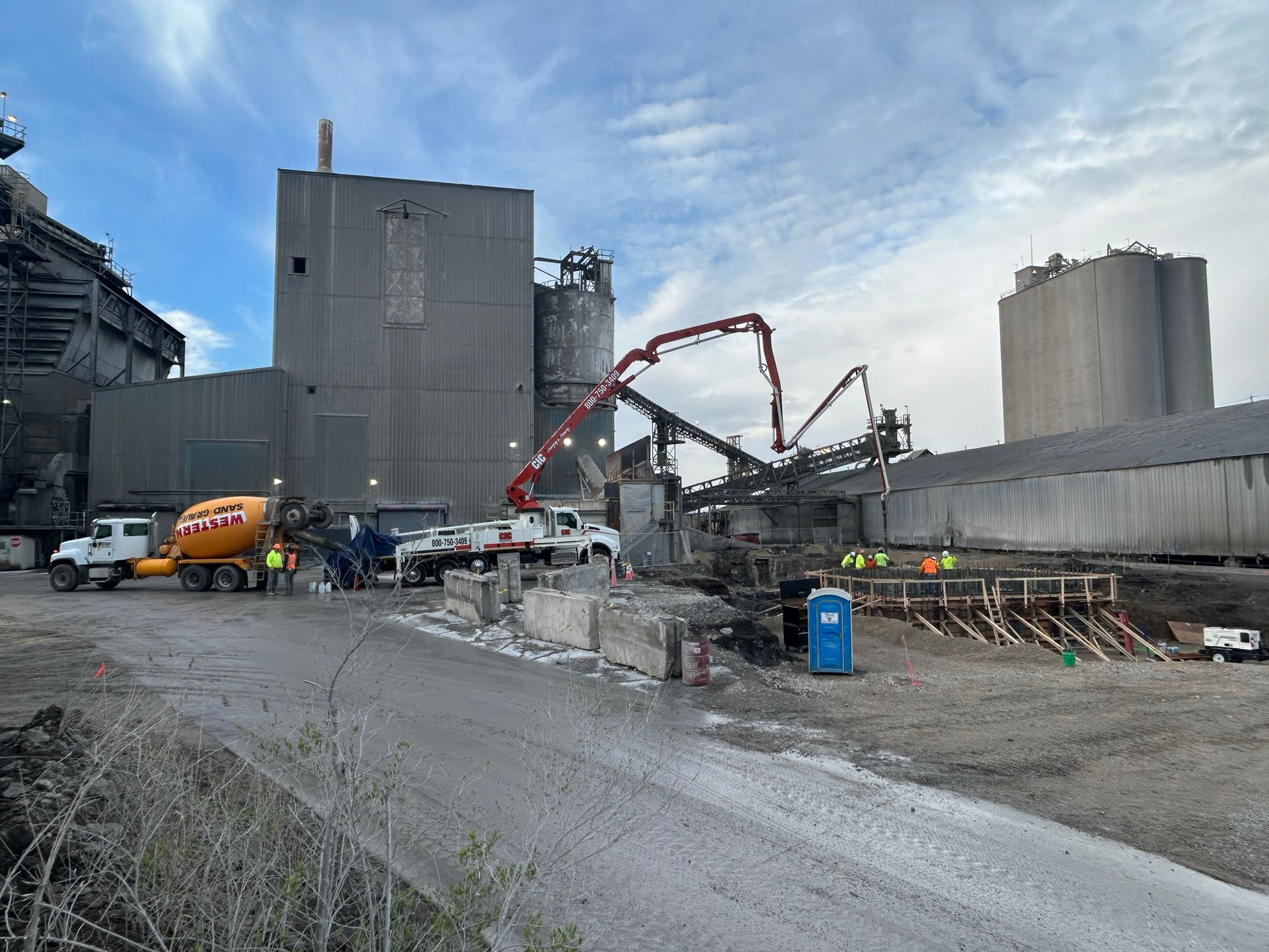 Construction site with cement trucks and machinery, pouring concrete near large industrial buildings under a cloudy sky.